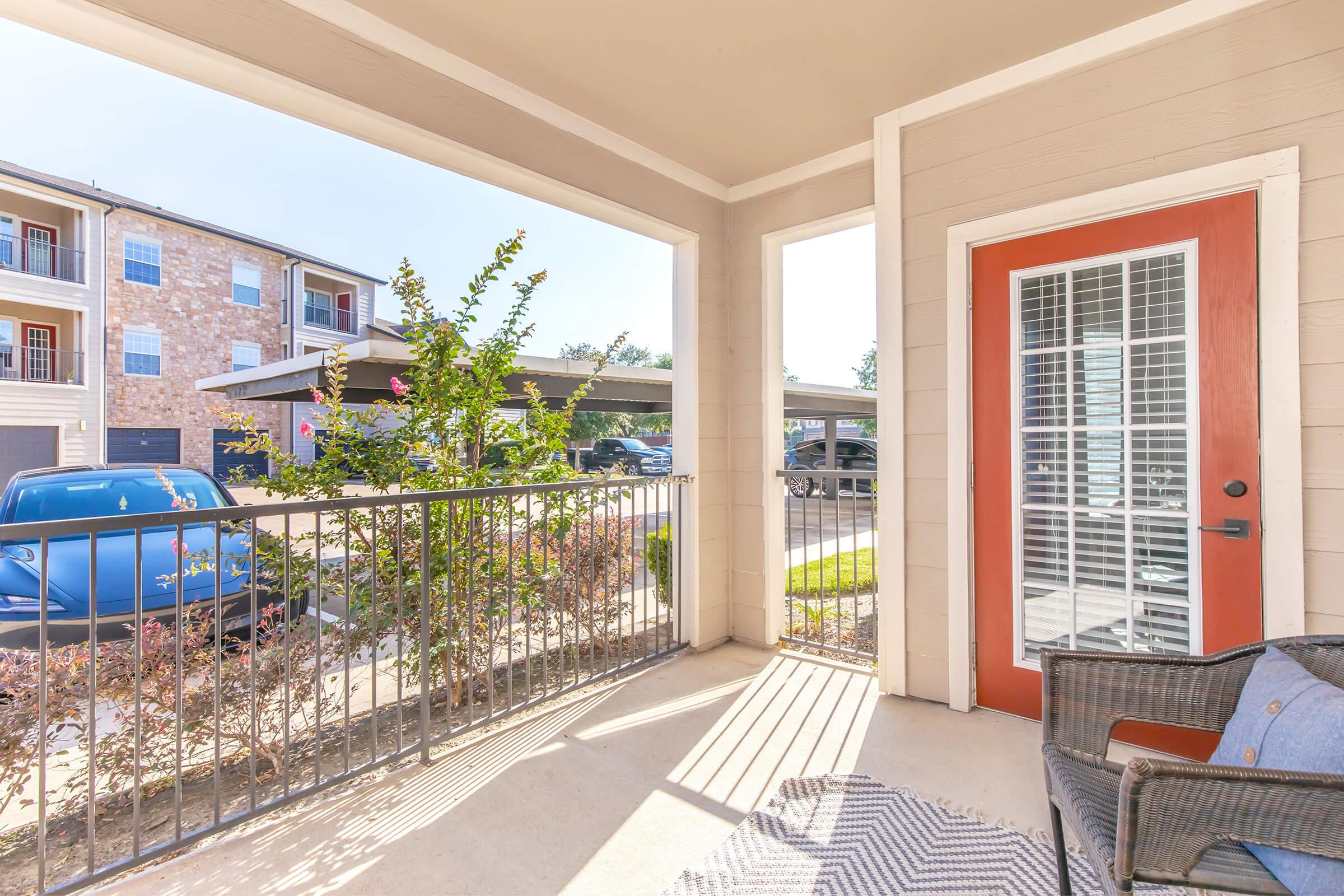 View from a covered patio showing a parked blue car and surrounding landscaping. A chair sits on the porch, with sunlight casting shadows on the floor. The scene includes apartment buildings in the background, highlighting a cozy outdoor space.