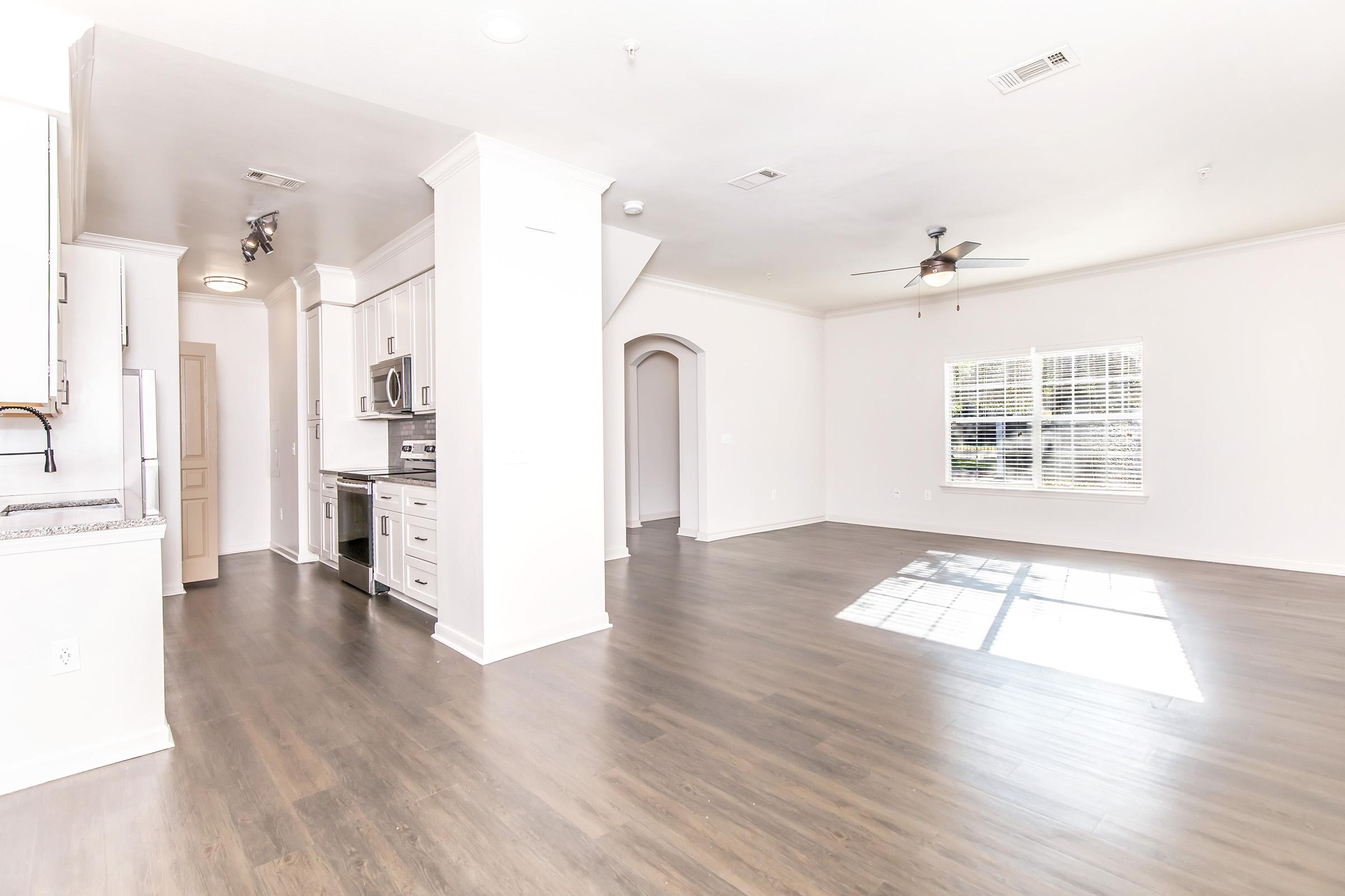 Spacious modern living area with an open layout, featuring light hardwood flooring and large windows allowing natural light. The kitchen includes stainless steel appliances and white cabinetry. A ceiling fan is present, and there’s an archway leading to another room.
