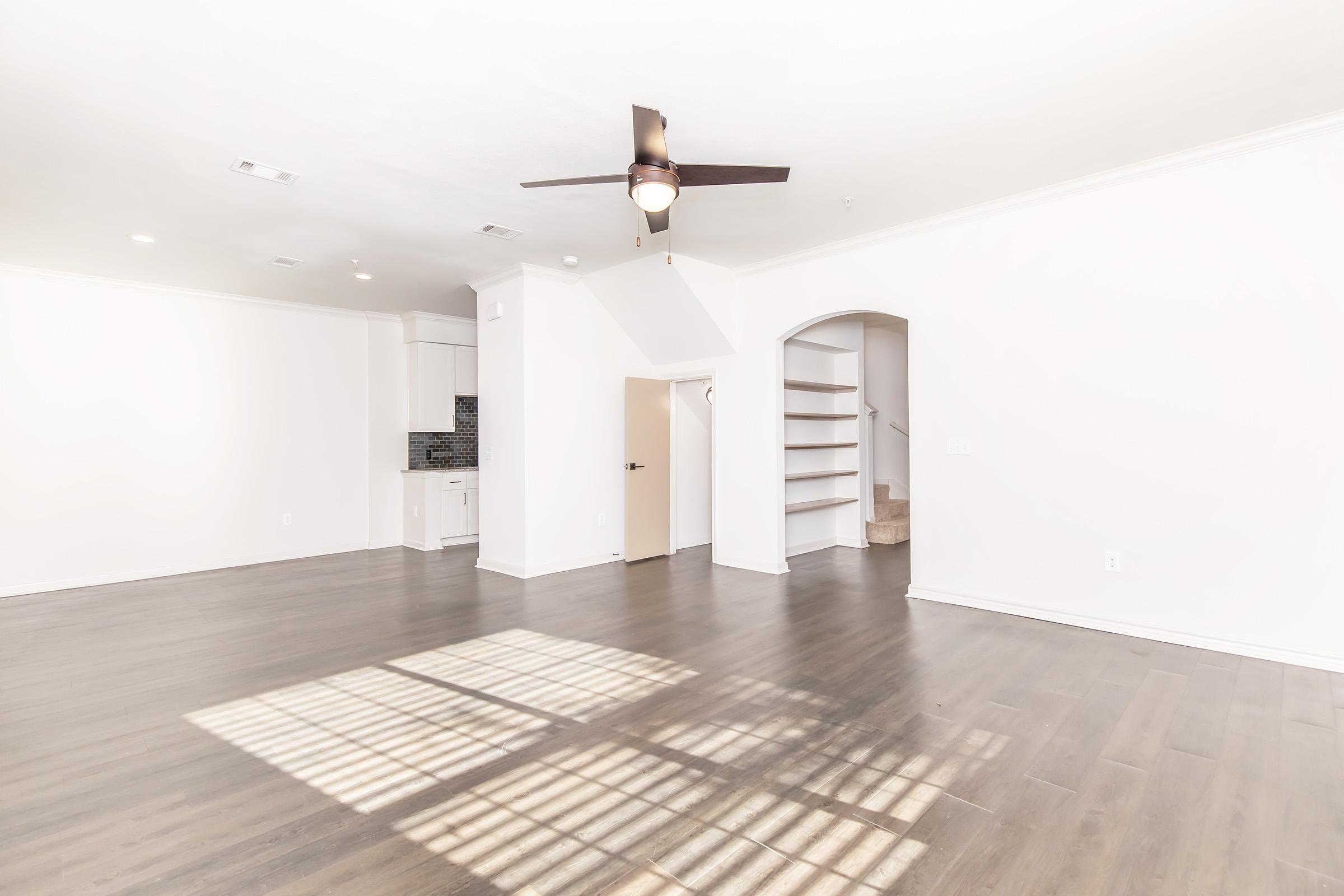 Spacious, well-lit living area featuring light wood flooring, a ceiling fan, and large windows casting shadows on the floor. To the right, an archway leads to a staircase, while the left side shows a modern kitchen with white cabinetry and a dark backsplash.