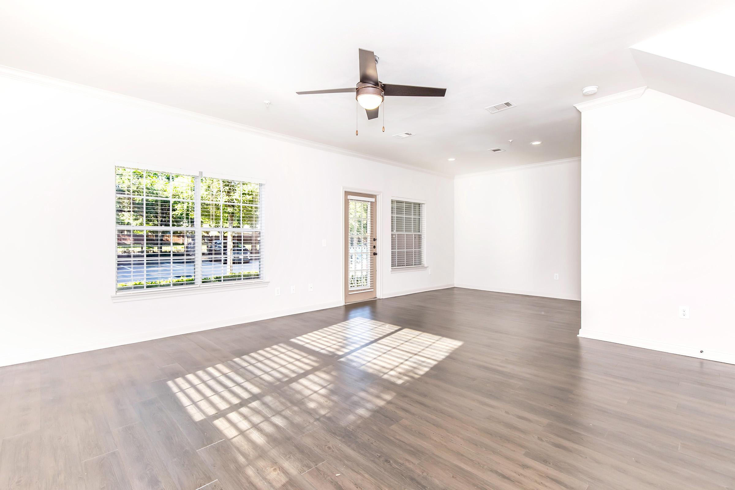 A spacious, empty living room with light-colored walls and dark hardwood flooring. A ceiling fan is mounted above, and large windows allow natural light to fill the space. A door leads to an outdoor area, and the room has a bright and airy feel.