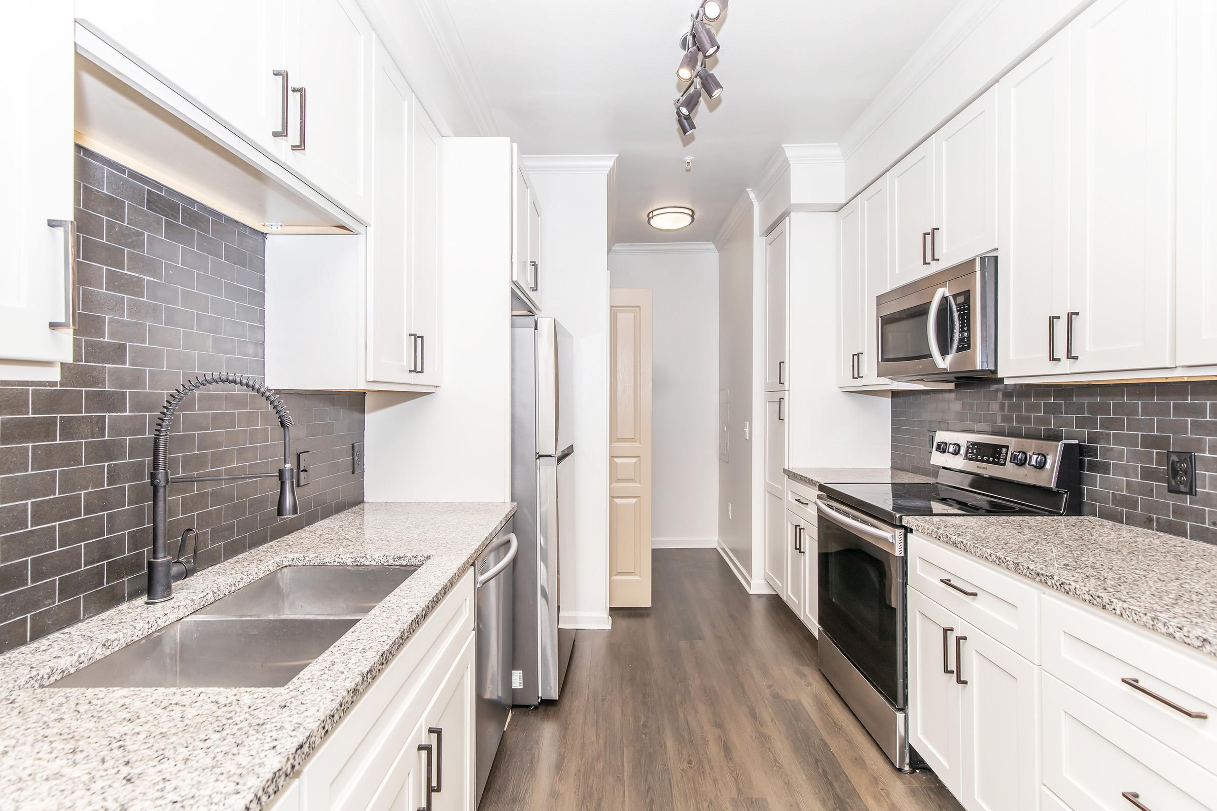 Modern kitchen featuring white cabinets, granite countertops, stainless steel appliances including a refrigerator, microwave, and oven. The backsplash is dark tiled, and there are recessed lights above. A dual sink with a sleek faucet is positioned under a window, creating a bright and functional space.