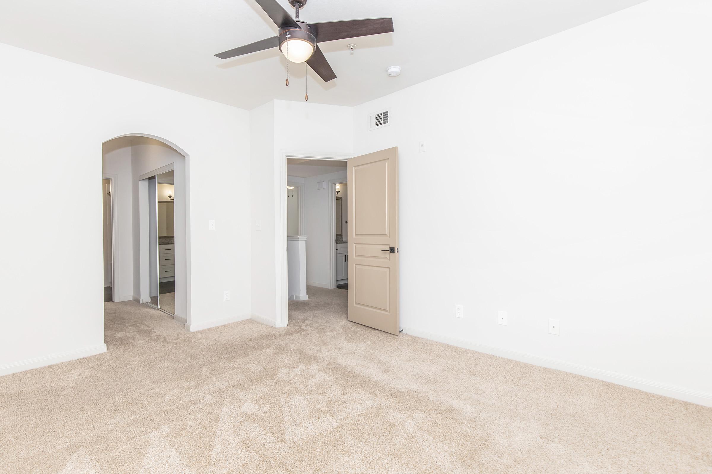 A spacious, empty bedroom with light beige carpet and plain white walls. A ceiling fan hangs from the ceiling, and there is an open door leading to a hallway. The room features an arched entrance on one side, with two mirrored closets visible in the background. Natural light fills the space.