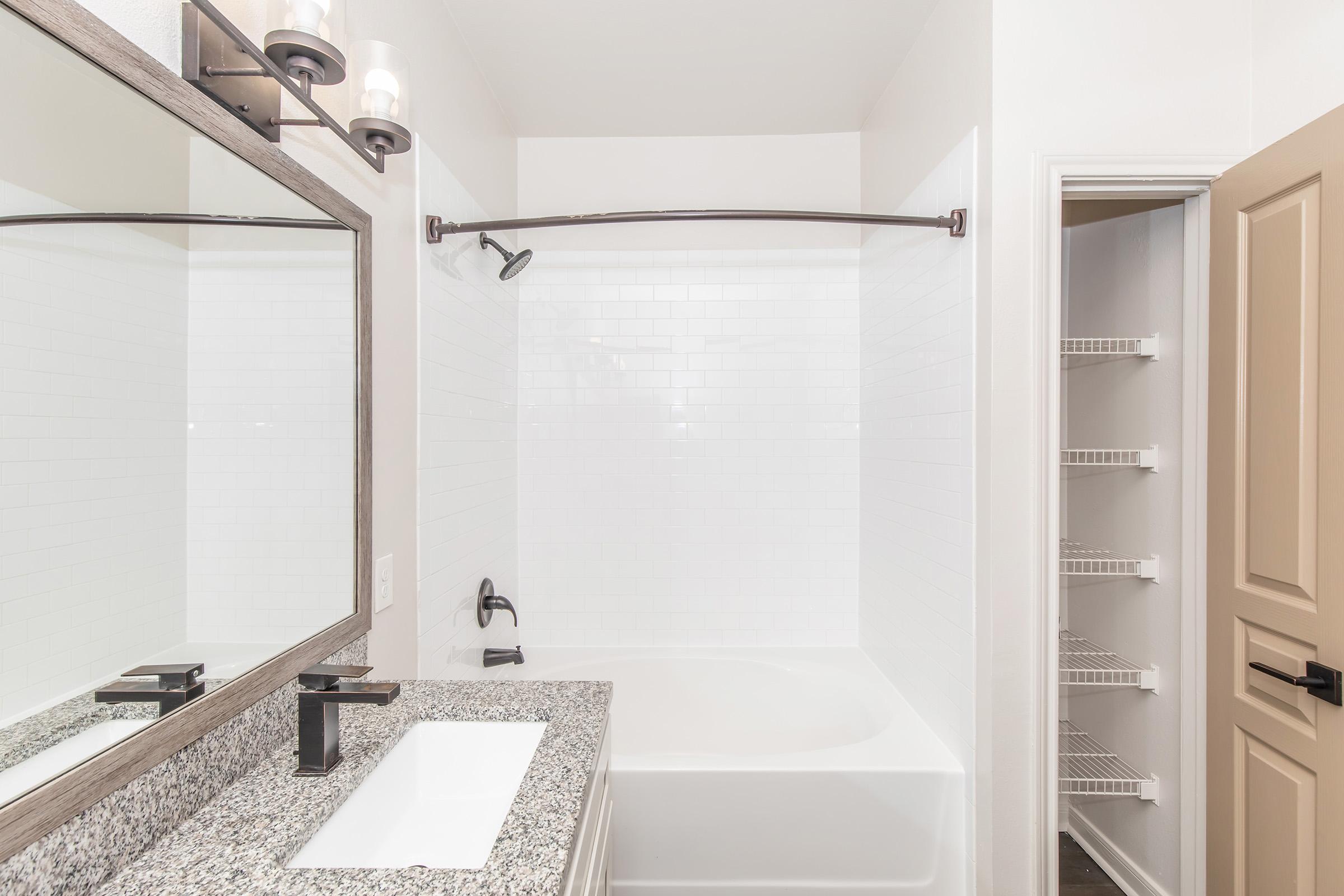 A modern bathroom featuring a bathtub with a shower, a large mirror above a granite countertop with a sink, and sleek black fixtures. To the right, there is an open door leading to a small pantry or storage area with white shelving units. The overall color scheme is light and contemporary.