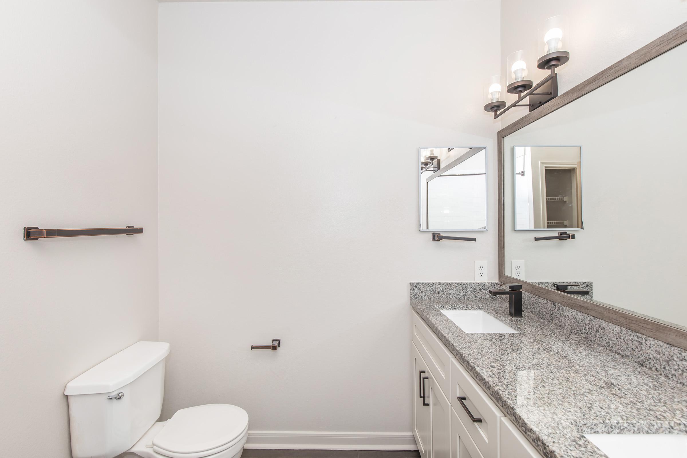 A clean, modern bathroom featuring a white toilet and a double vanity with a granite countertop. Above the vanity are two square mirrors and three wall-mounted light fixtures. The walls are a neutral tone, and the floor is a dark laminate, creating a contemporary and spacious atmosphere.