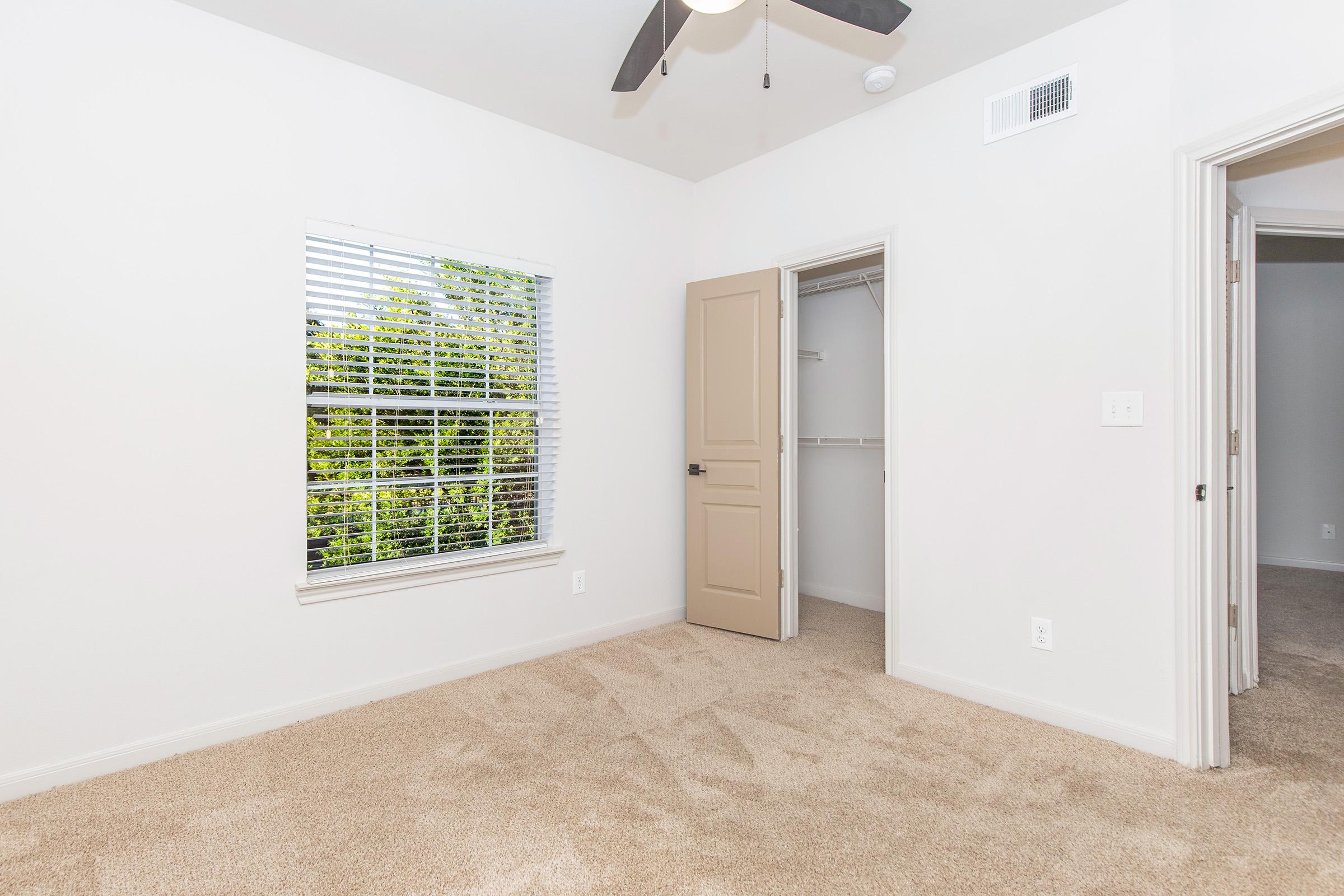 A well-lit room with beige carpet, featuring a ceiling fan and white walls. There is a window with blinds allowing natural light, and a closet visible through an open door. The room appears empty and ready for furniture, with another door leading to an adjacent space.