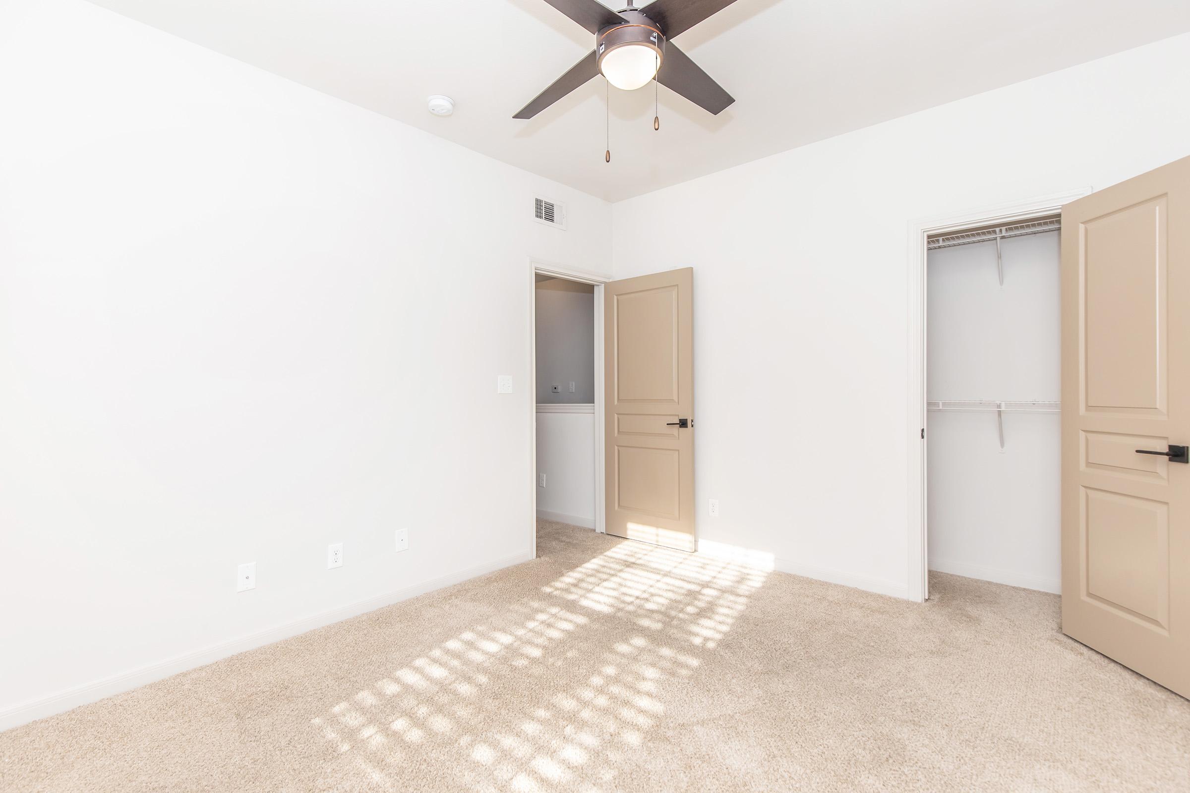 A well-lit empty room with light-colored walls and carpet. It features a ceiling fan, two doors—one leading to a closet and another possibly to a bathroom or hallway. Sunlight casts decorative shadows on the floor, enhancing the spacious feel of the area.