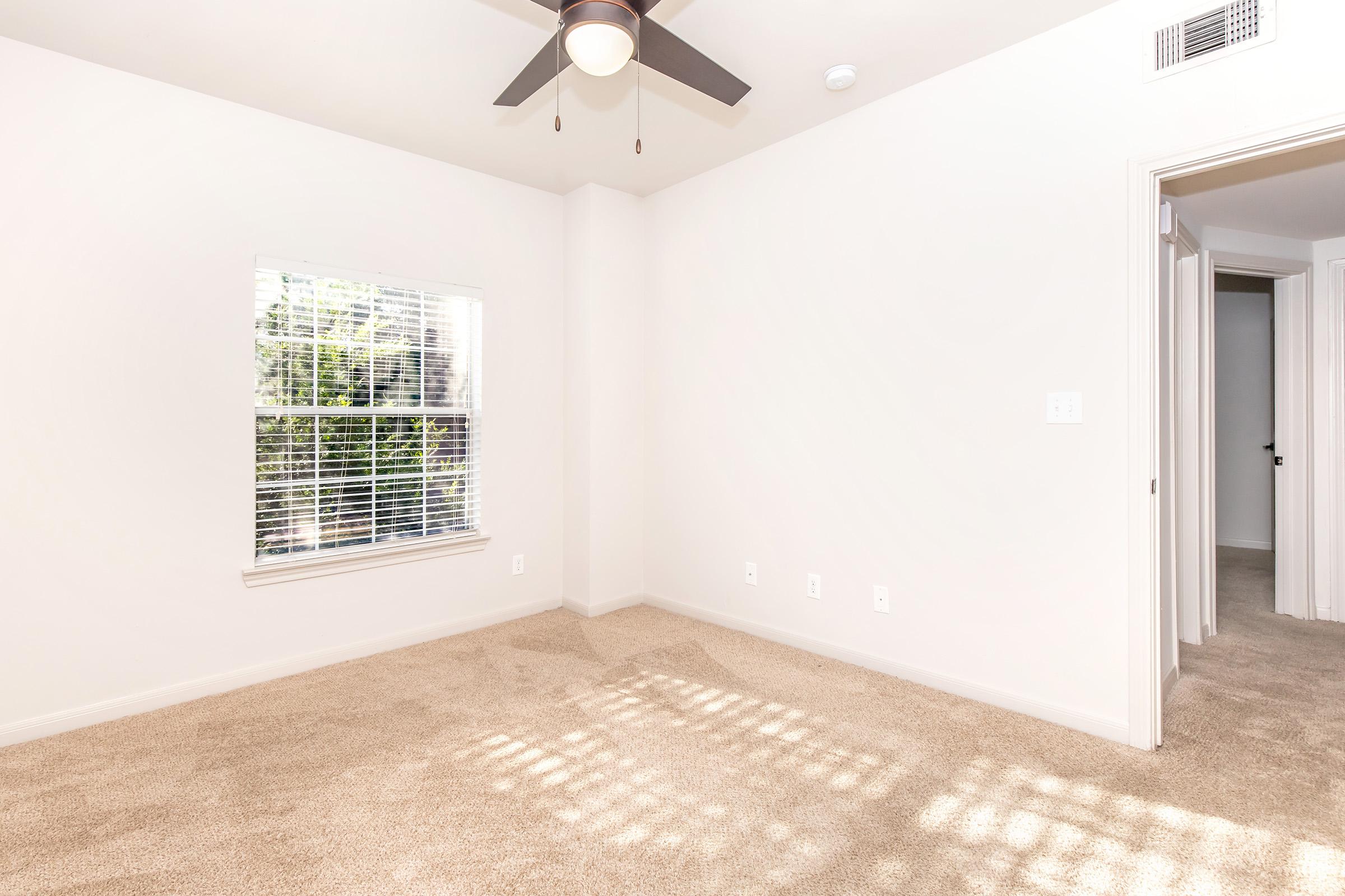 A bright, empty room featuring light beige carpet, a ceiling fan, and a window with white blinds allowing natural light. The walls are painted in a soft color, creating a clean and airy atmosphere. There are doorways leading to other rooms, emphasizing the spacious layout.