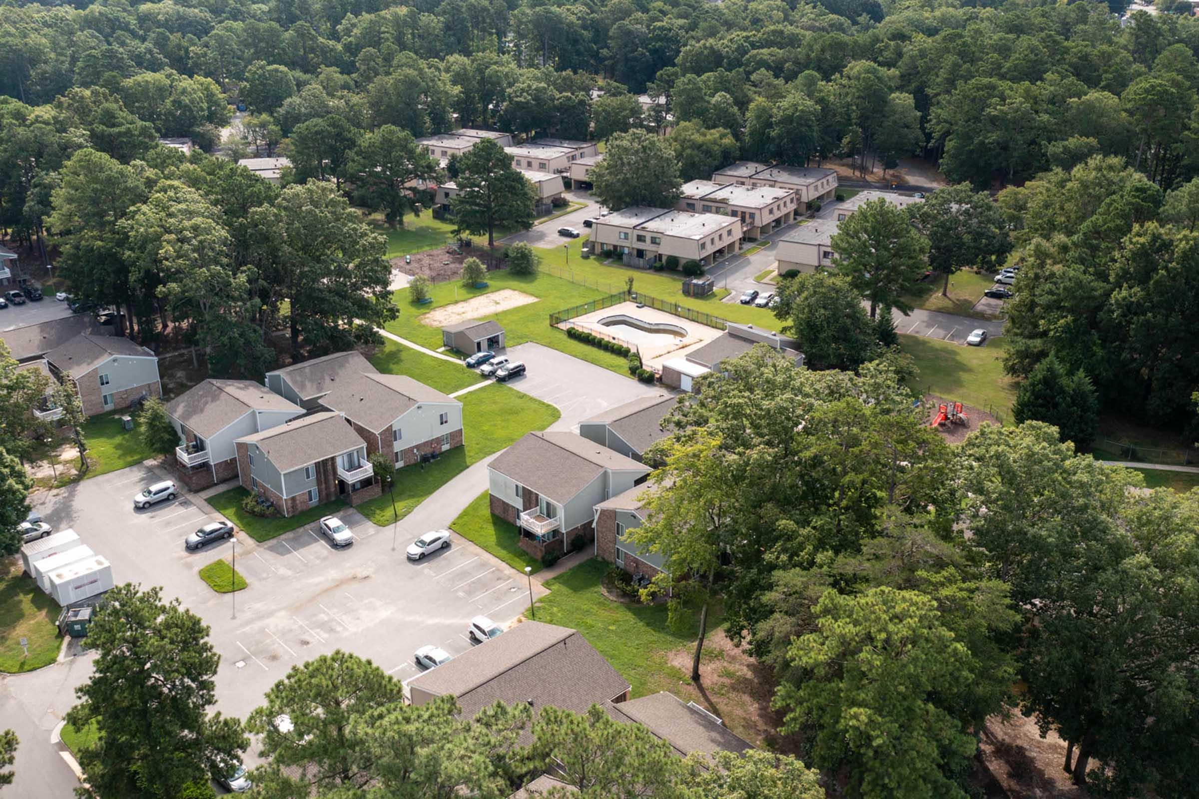 Aerial view of a residential area featuring several two-story apartment buildings surrounded by trees and grassy lawns. Some vehicles are parked in the lot, and a community area with a swimming pool is visible. Nearby, there are playgrounds and additional housing structures among the greenery.