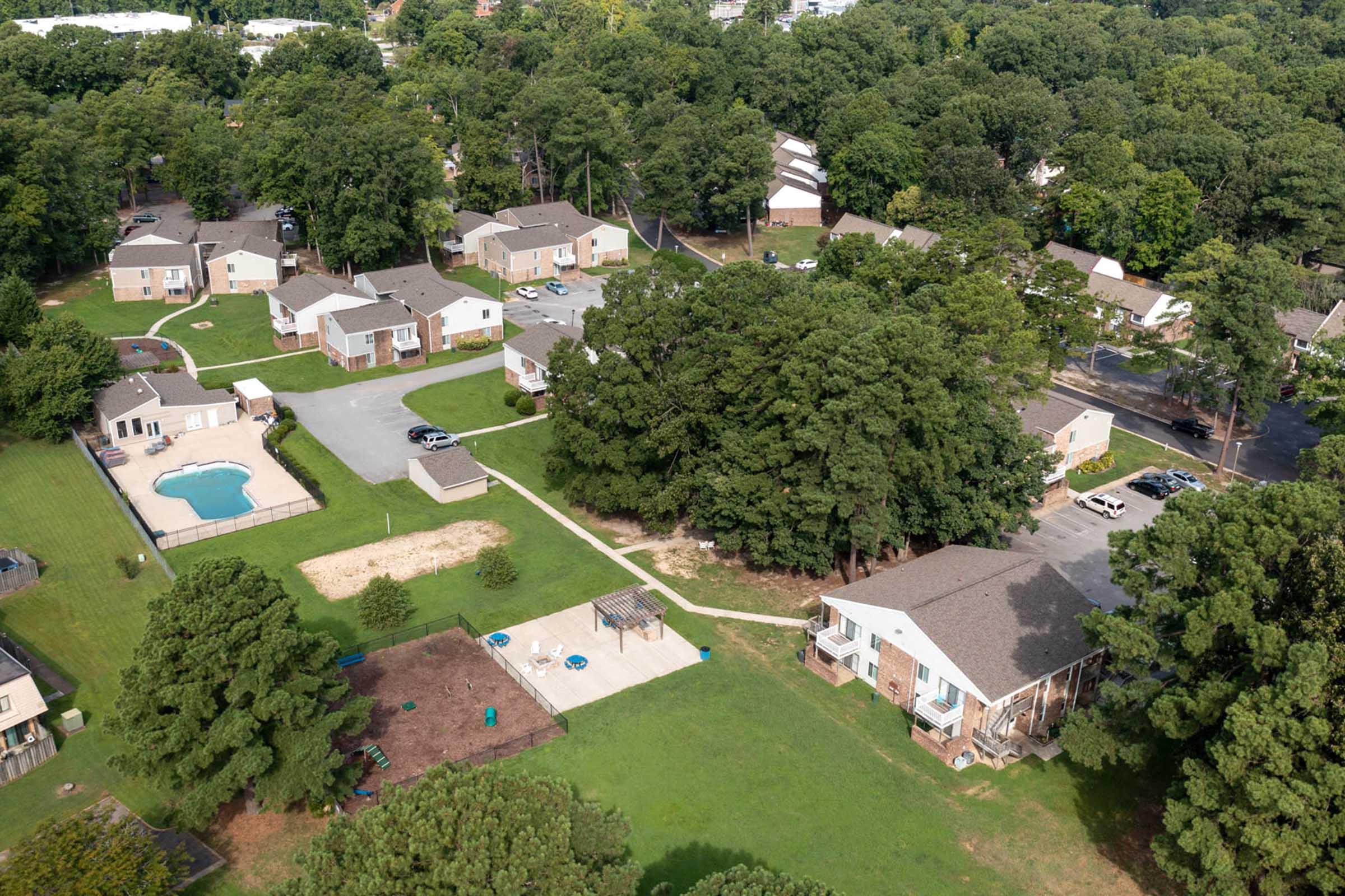 Aerial view of a residential area featuring several apartment buildings surrounded by green trees. A multi-use playground and a swimming pool are visible, along with grassy areas and parking spaces. The scene conveys a peaceful suburban setting with ample outdoor space.