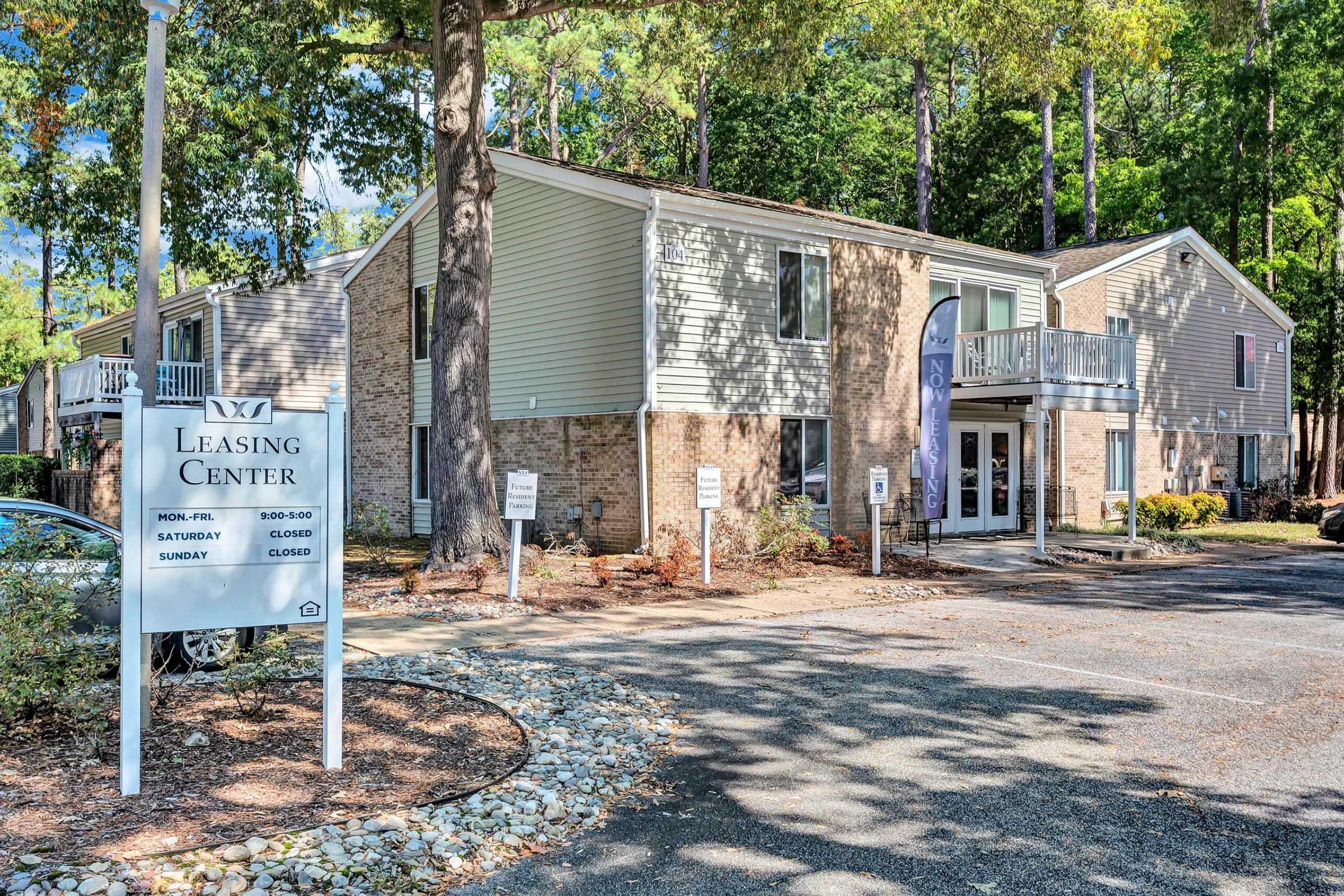Leasing center building in a residential area, featuring a sign with operating hours. The structure has multiple windows and a small balcony. Surrounding the building are trees and landscaped areas, with clear signage indicating the leasing office is open Monday to Friday, and closed on weekends.