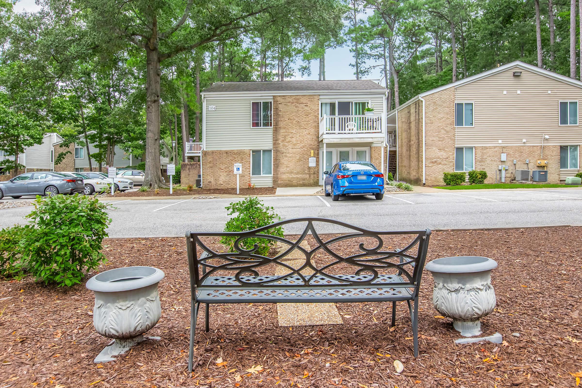 A metal bench with decorative designs is positioned in front of two multi-unit residential buildings surrounded by trees. There are potted plants on either side of the bench, and a blue car is parked nearby in the lot. The area has a neat, well-maintained appearance with mulch covering the ground.