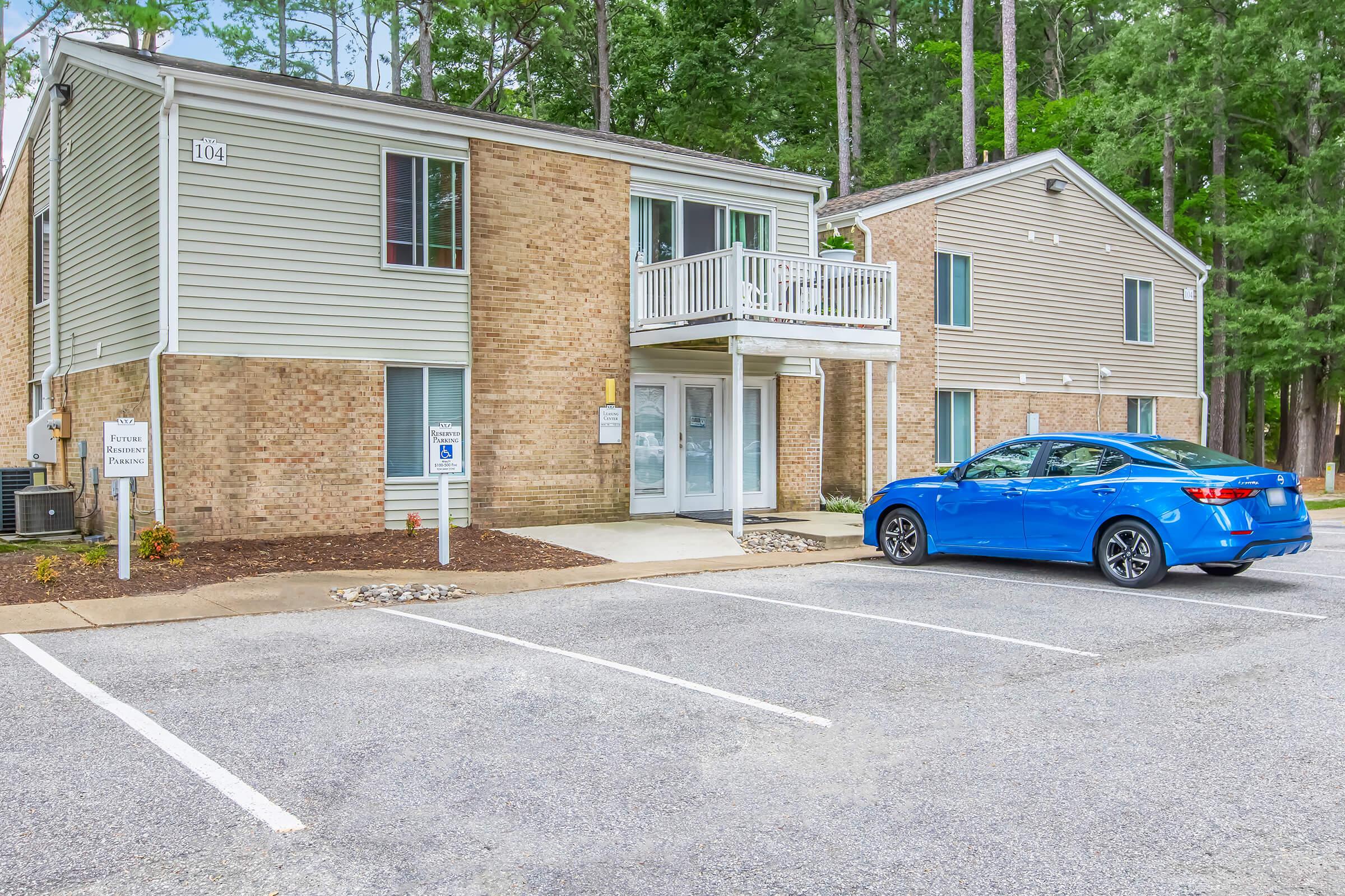 Two multi-story apartment buildings with a brick facade and light-colored vinyl siding. The front entrance of the building is clearly visible, featuring a balcony above. A blue car is parked in front, with a parking lot and trees in the background creating a serene atmosphere.