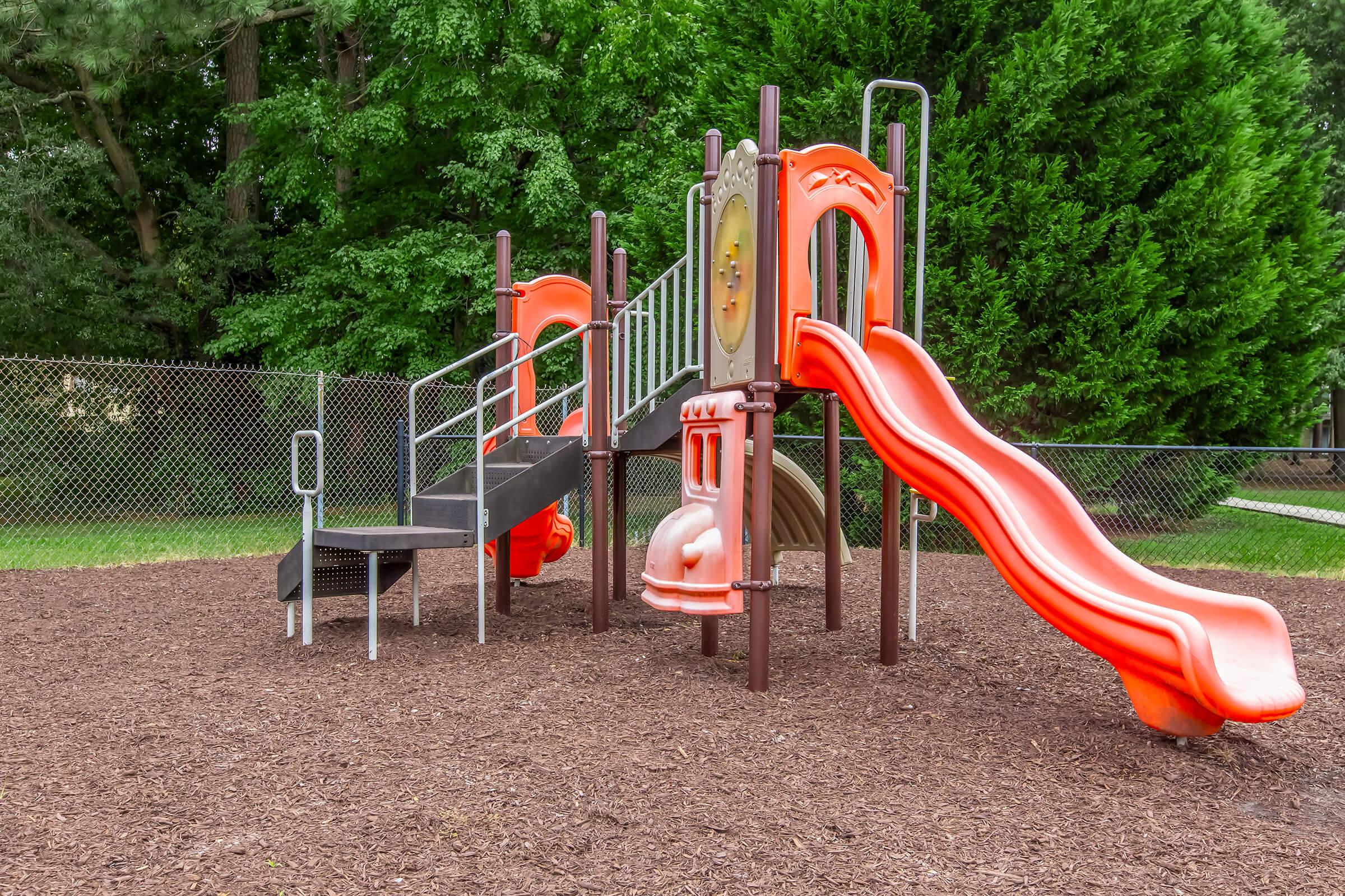 A colorful playground featuring a slide and climbing structure, set on a bed of wood chips. The equipment is orange and gray, surrounded by green trees and a chain-link fence. Ideal for children to play and explore.