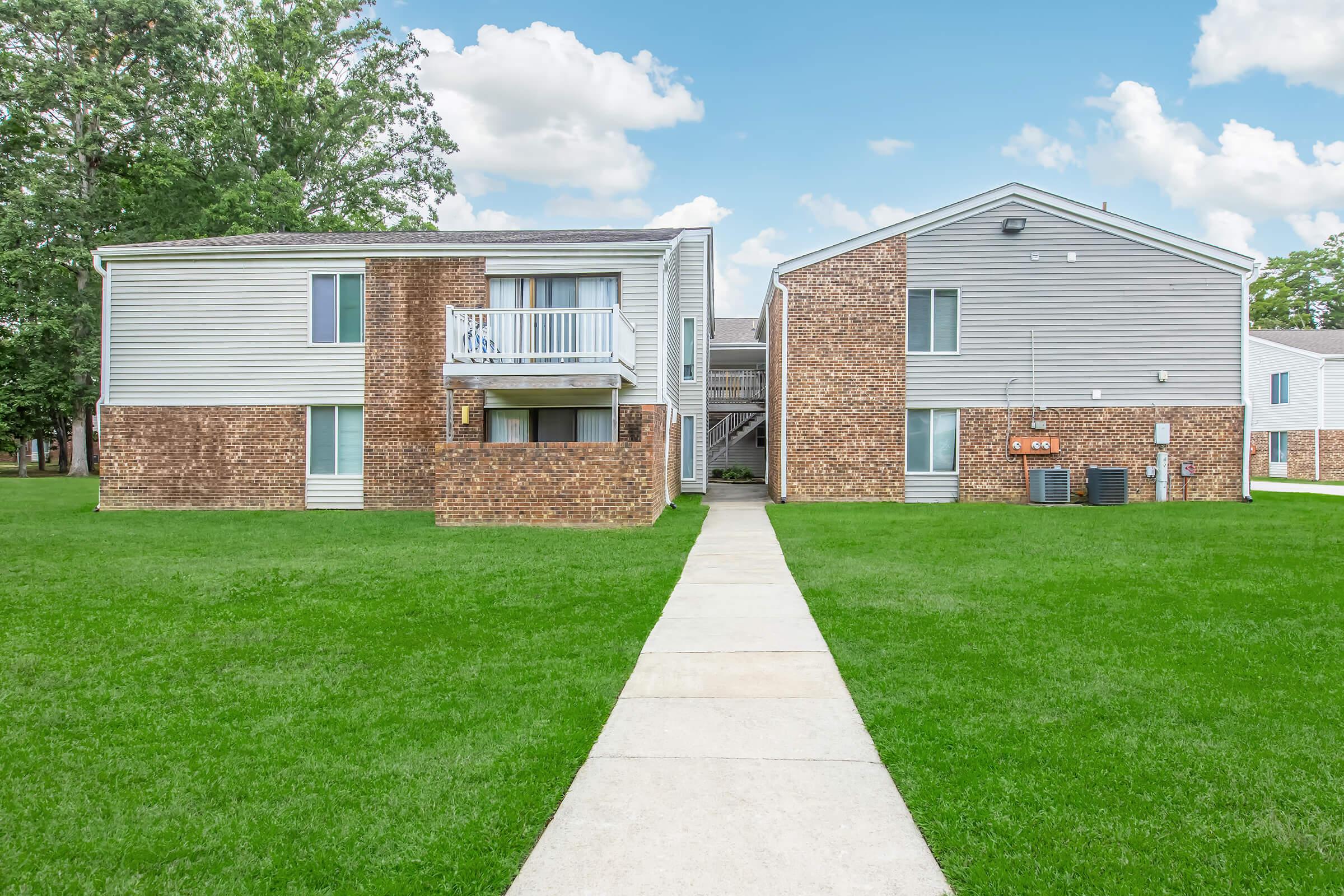 Two multi-unit residential buildings with a combination of gray siding and brick facades, surrounded by well-maintained green grass. A walkway leads through the grass, connecting the two buildings. The sky is bright with scattered clouds.