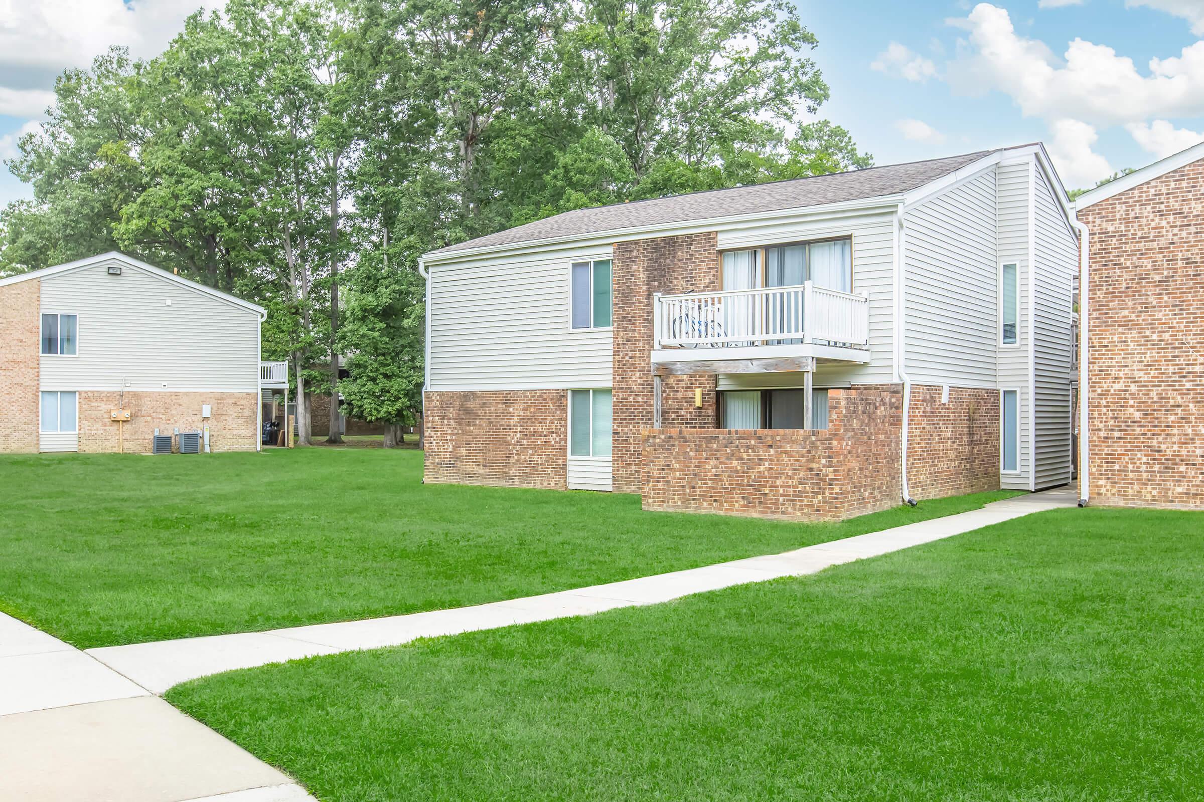 A two-story apartment building featuring a mix of light-colored siding and brick exterior. The grass lawn is green and well-maintained, with a clear pathway leading to the entrance. In the background, another similar building can be seen, surrounded by trees under a partly cloudy sky.