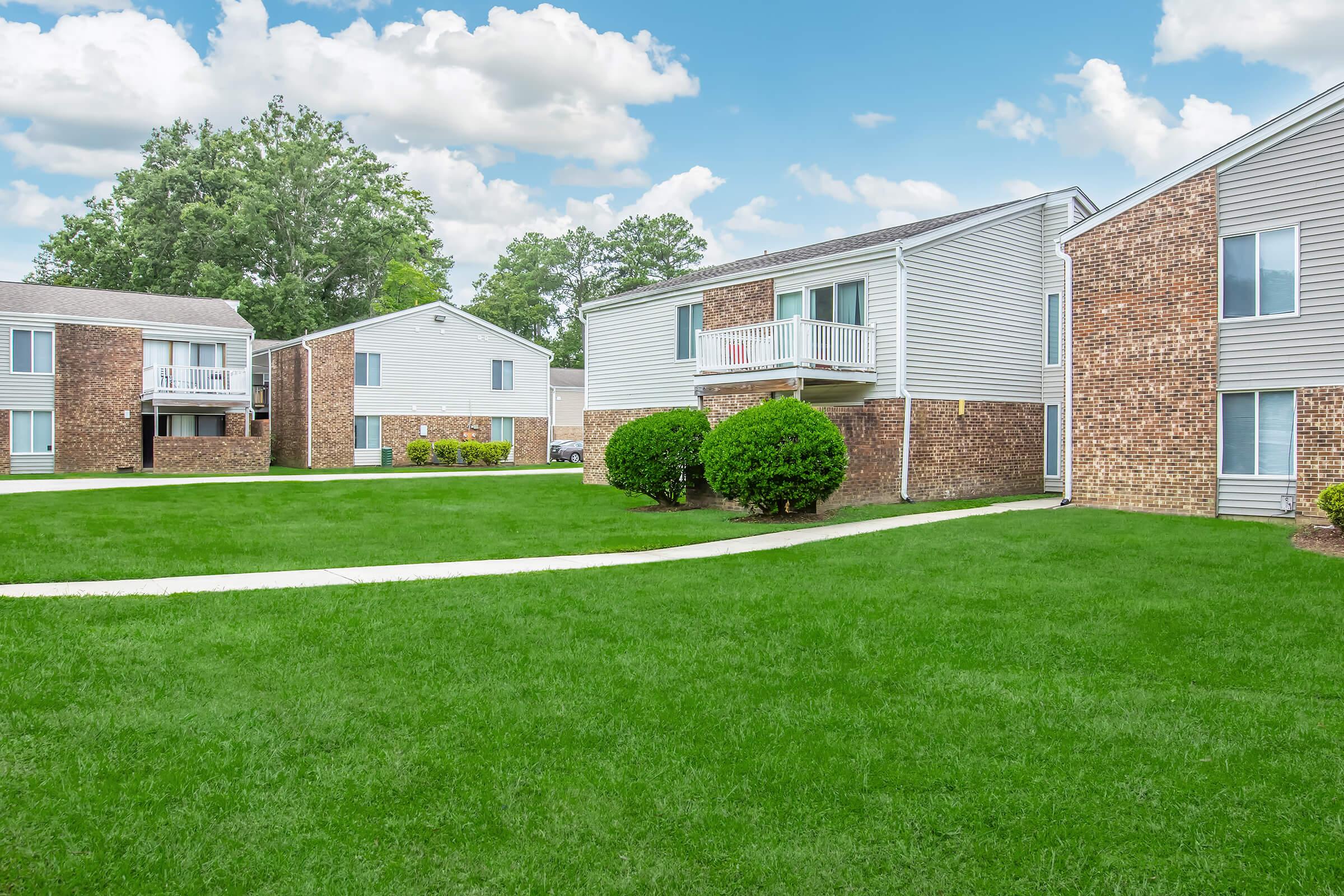 A well-maintained apartment complex featuring several brick and siding buildings. The landscape includes a green lawn and neatly trimmed bushes, with a clear blue sky and fluffy white clouds in the background. Pathways lead between the buildings, creating an inviting residential atmosphere.