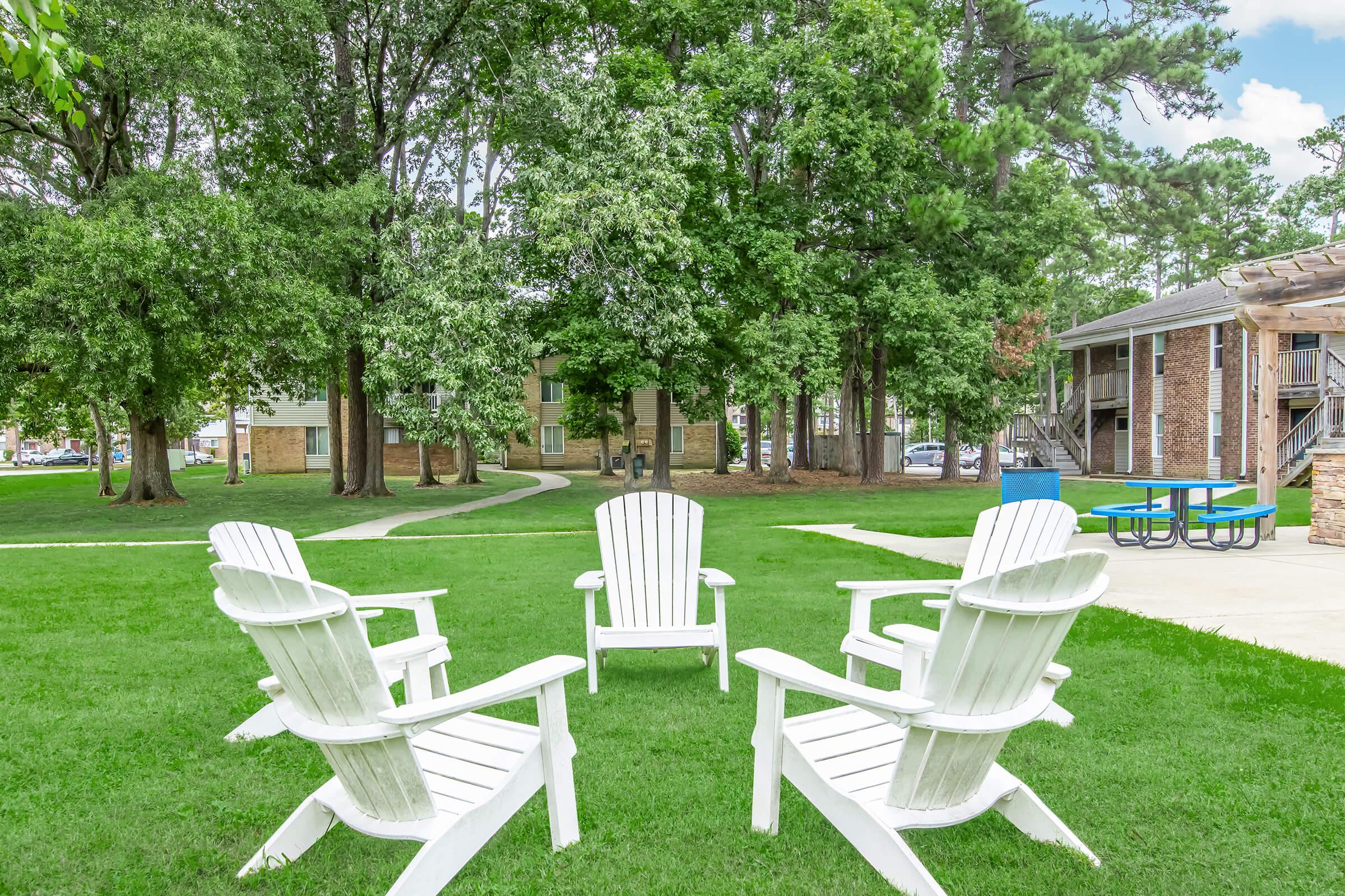 A serene outdoor setting featuring four white Adirondack chairs arranged in a circle on lush green grass, with trees in the background and an apartment building visible nearby. The scene invites relaxation and social gatherings in a pleasant environment.