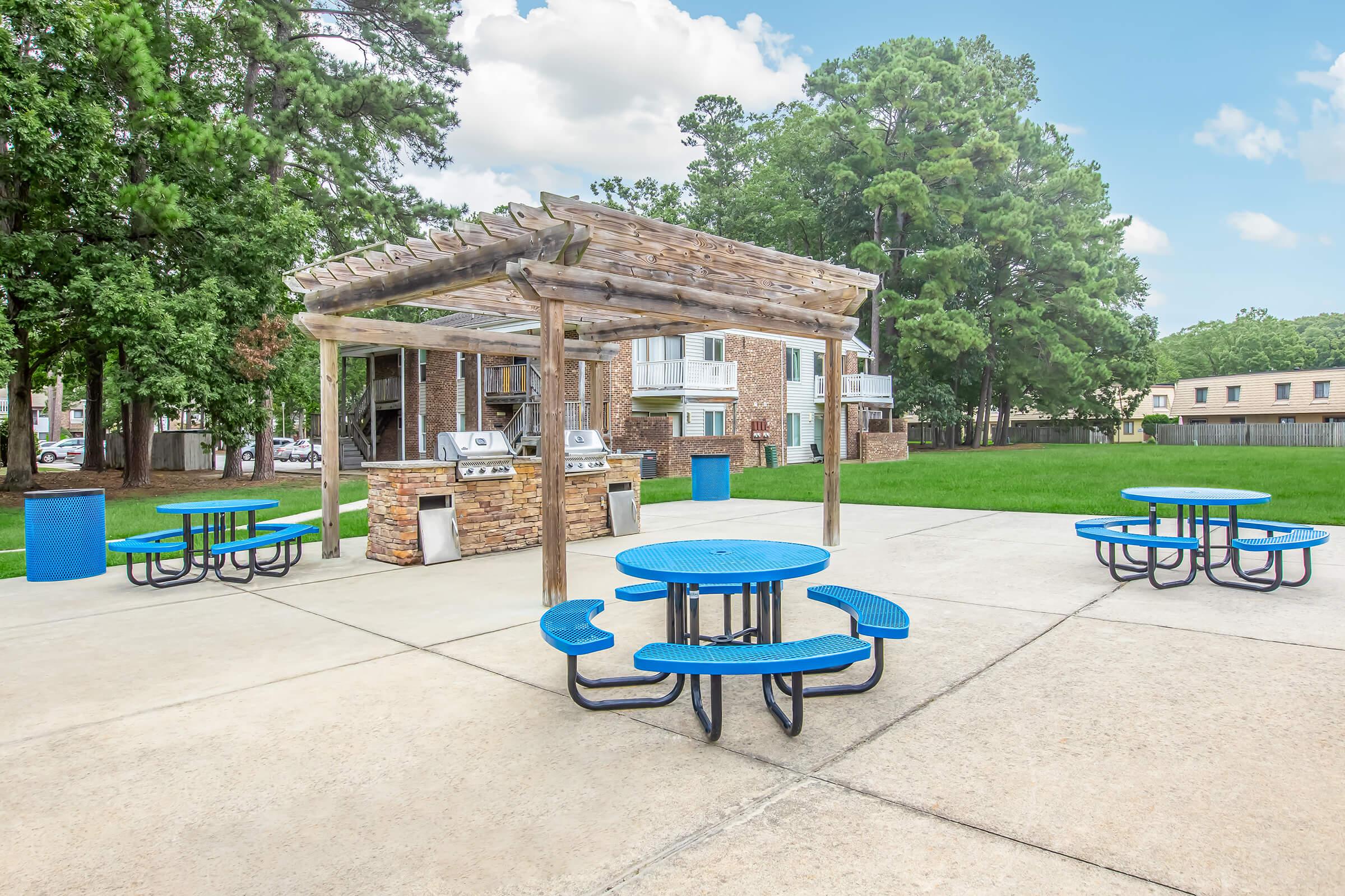 Outdoor recreational area featuring blue picnic tables and a stone grill station under a wooden pergola. The space is surrounded by greenery and residential buildings in the background, with a clear sky above. The setting is ideal for gatherings and barbecues.