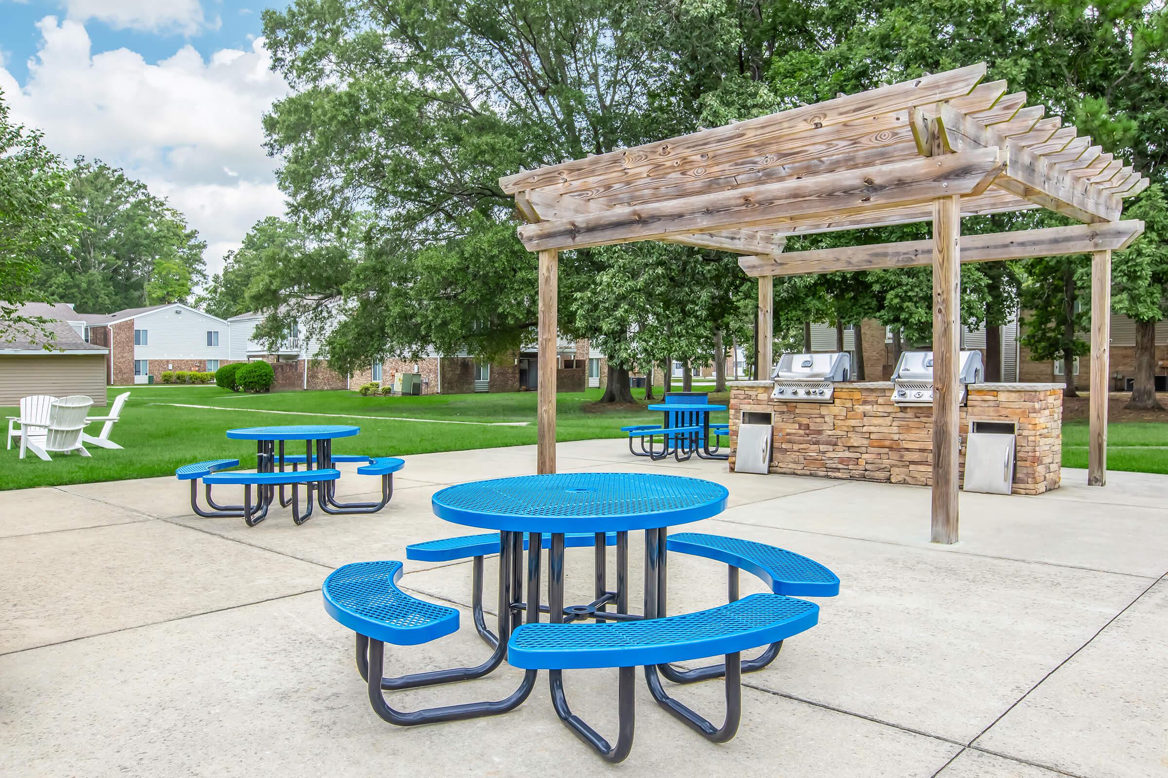 An outdoor area featuring blue picnic tables arranged around a concrete patio, with a shaded pergola overhead. Two barbecue grills are positioned beneath the pergola, surrounded by green lawns and residential buildings in the background. The scene is bright and inviting, perfect for gatherings.
