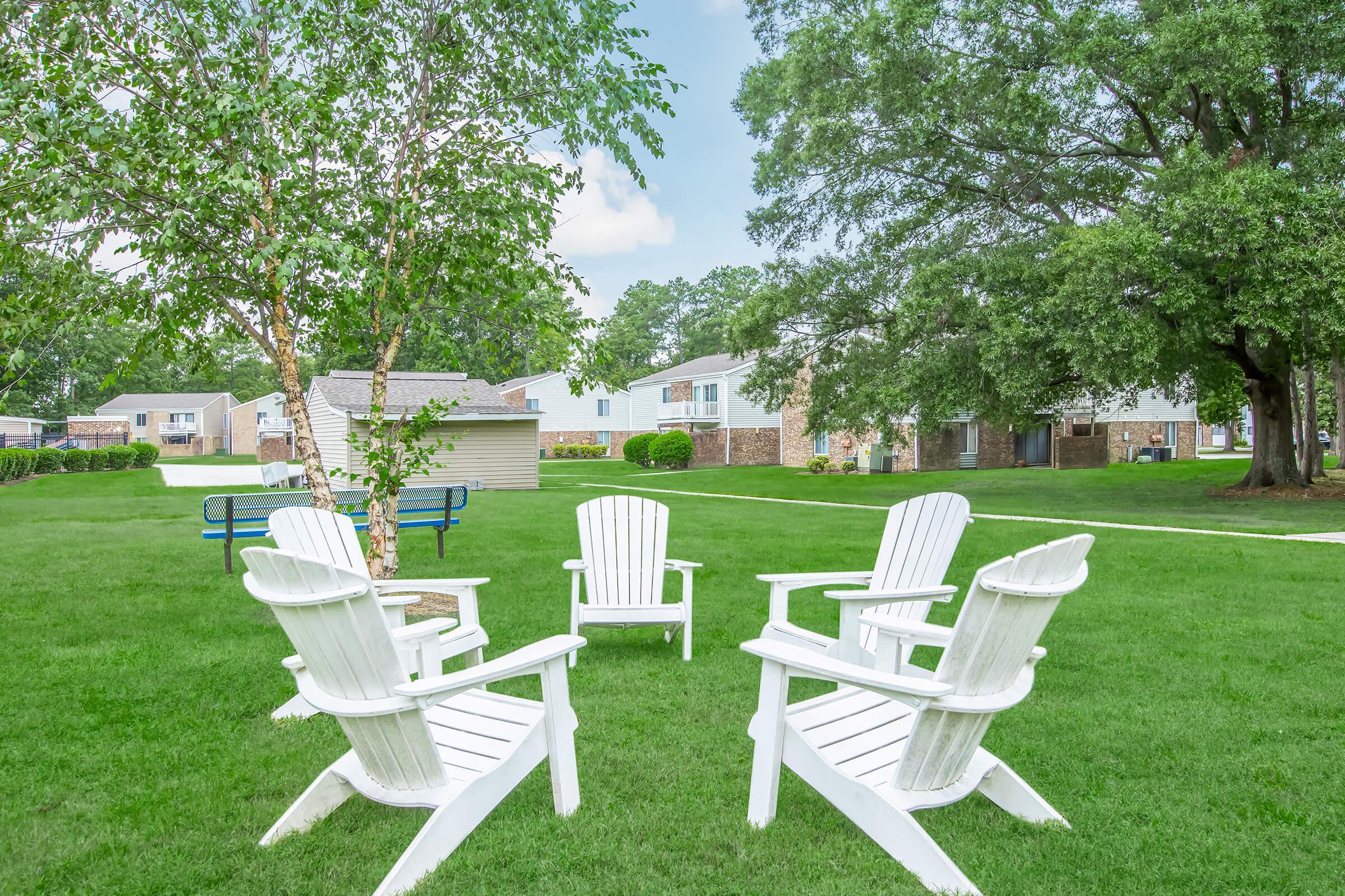 A peaceful outdoor scene featuring four white Adirondack chairs arranged in a circular formation on a lush green lawn. In the background, there are trees and residential buildings, creating a serene atmosphere perfect for relaxation or gathering.