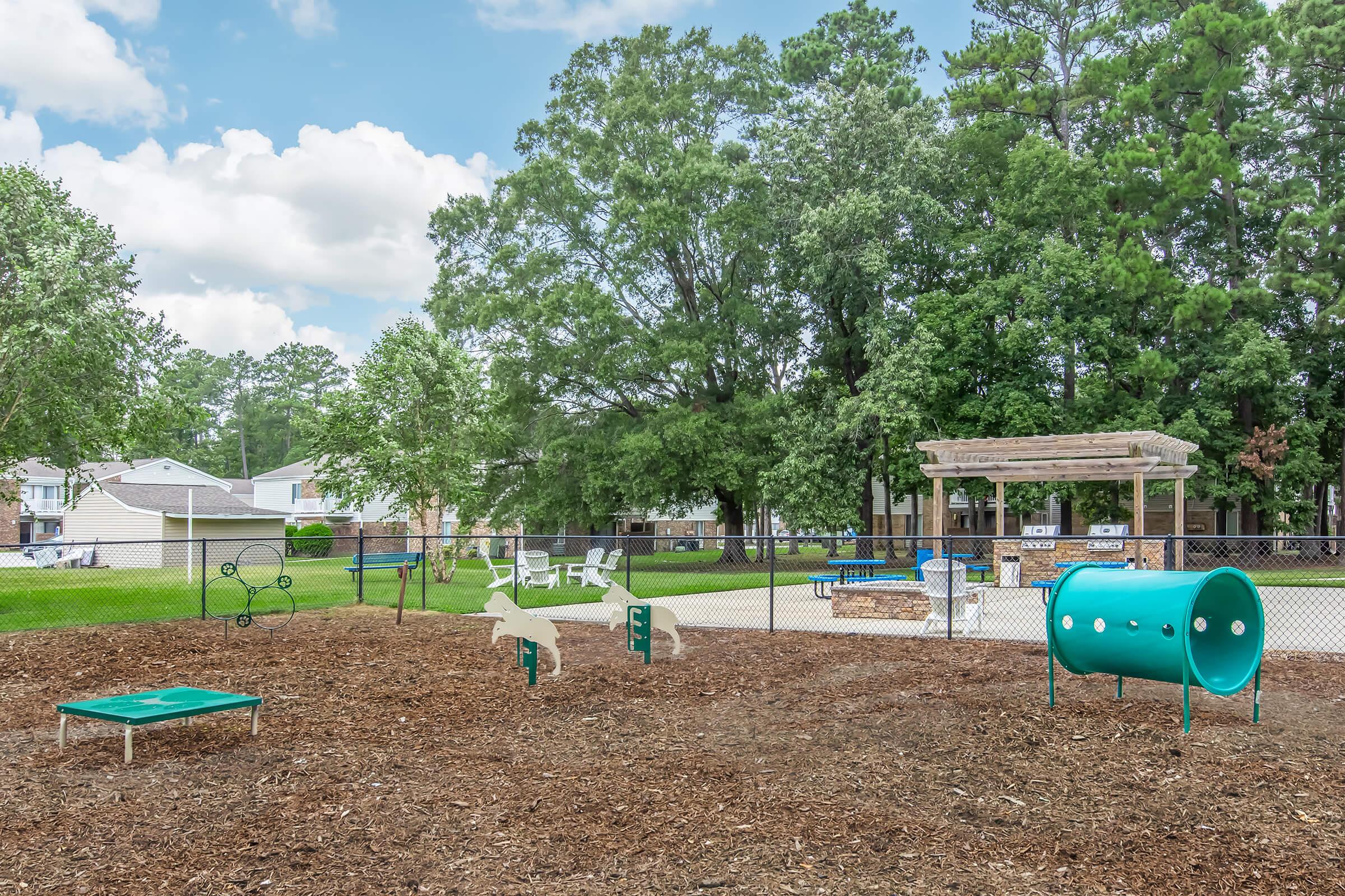 A spacious dog park featuring agility equipment like a tunnel, jumps, and climbing platforms. Surrounded by lush trees and green grass, there are picnic tables and benches nearby. The park is enclosed with a fence, creating a safe area for dogs to play and socialize.