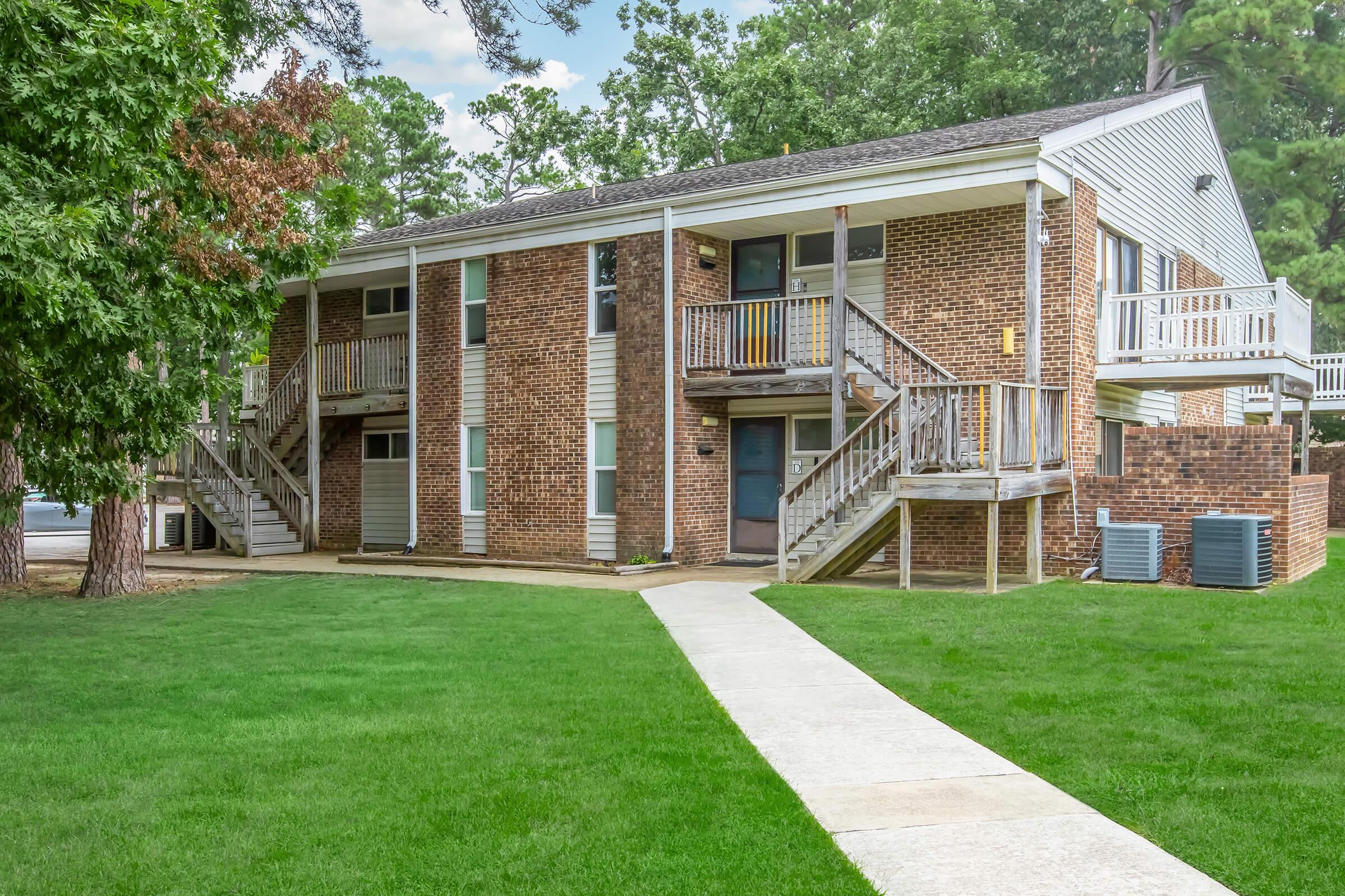 A two-story brick apartment building with balconies, surrounded by green grass and trees. A concrete pathway leads to the entrance. Air conditioning units are visible near the building's base. The sky is partly cloudy.