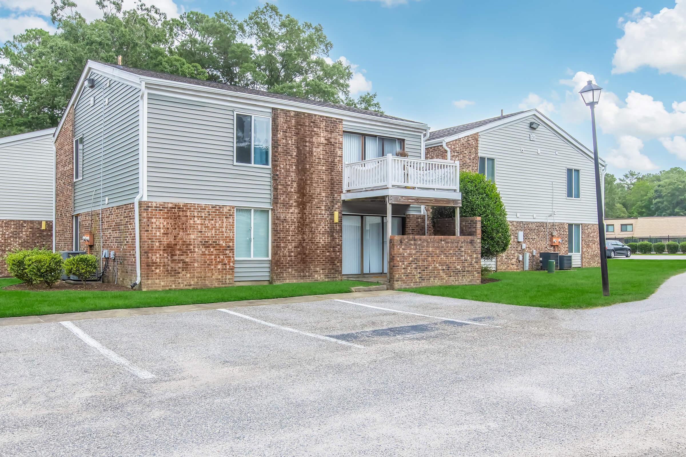 A view of a two-story apartment complex featuring a mix of brick and siding exteriors. The foreground includes a parking area with several empty spaces, and green lawns surround the buildings. A lamppost stands in the driveway, and the sky is partly cloudy.