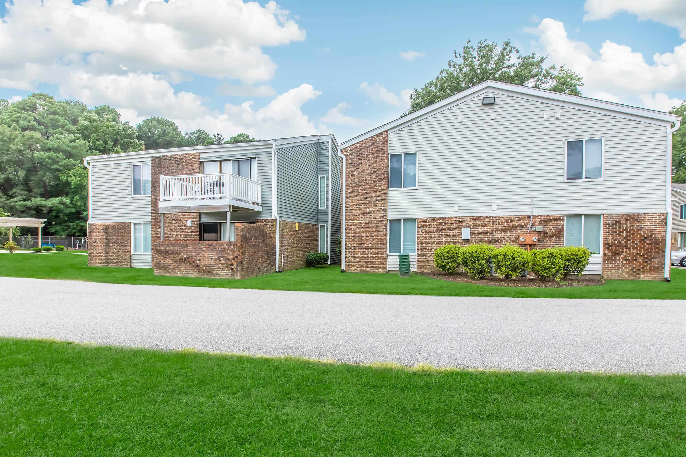 Two multi-unit residential buildings made of brick and siding, situated on a grassy area with a gravel road in front. The buildings have large windows and some balconies. Surrounding greenery includes bushes and trees, with a cloudy blue sky above.