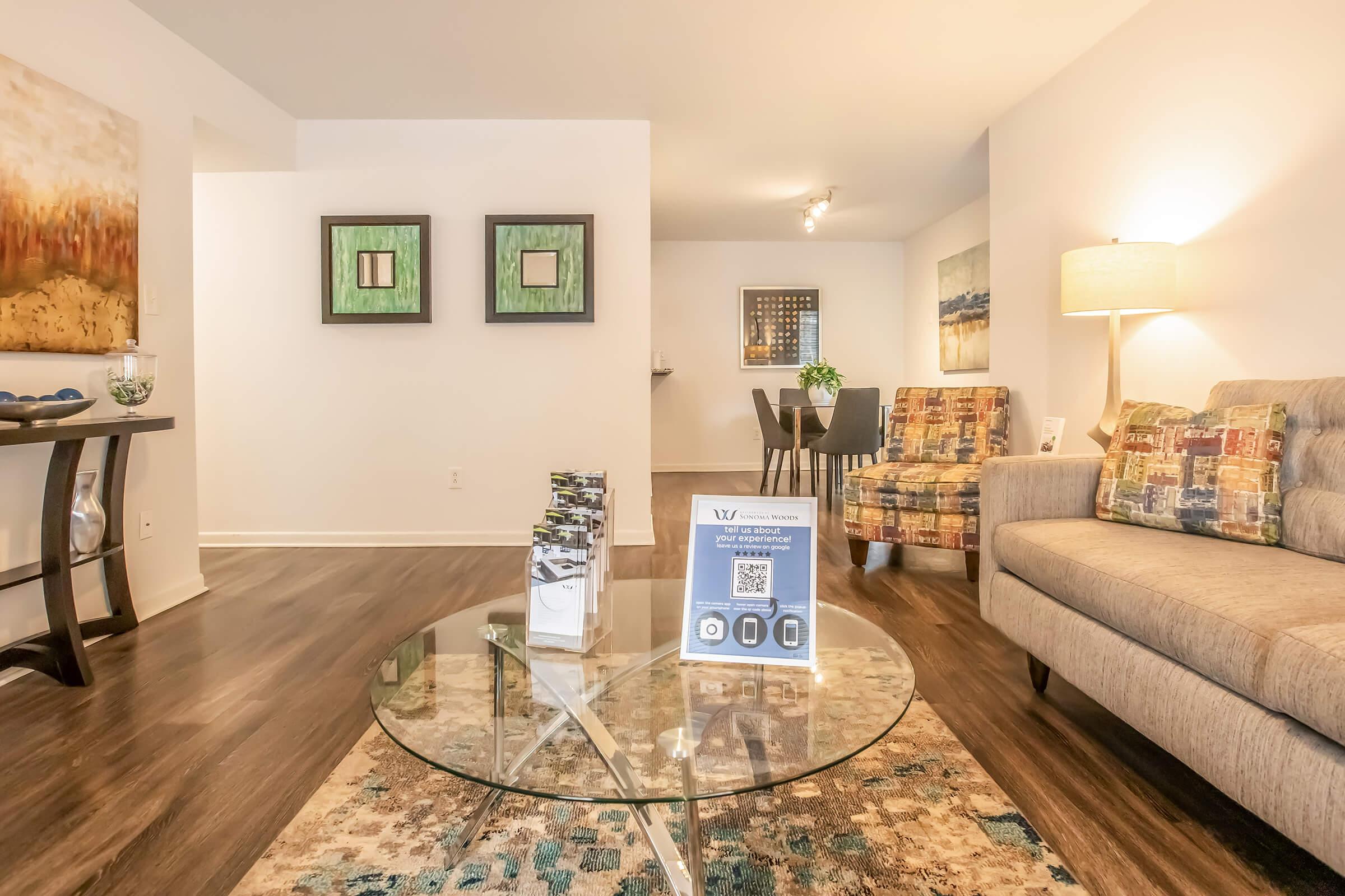 A modern living room featuring a light-colored couch with patterned cushions, a glass coffee table with brochures, and decorative wall art. The space includes a small dining area with a table and chairs, hardwood floors, and a warm lamp providing soft lighting.