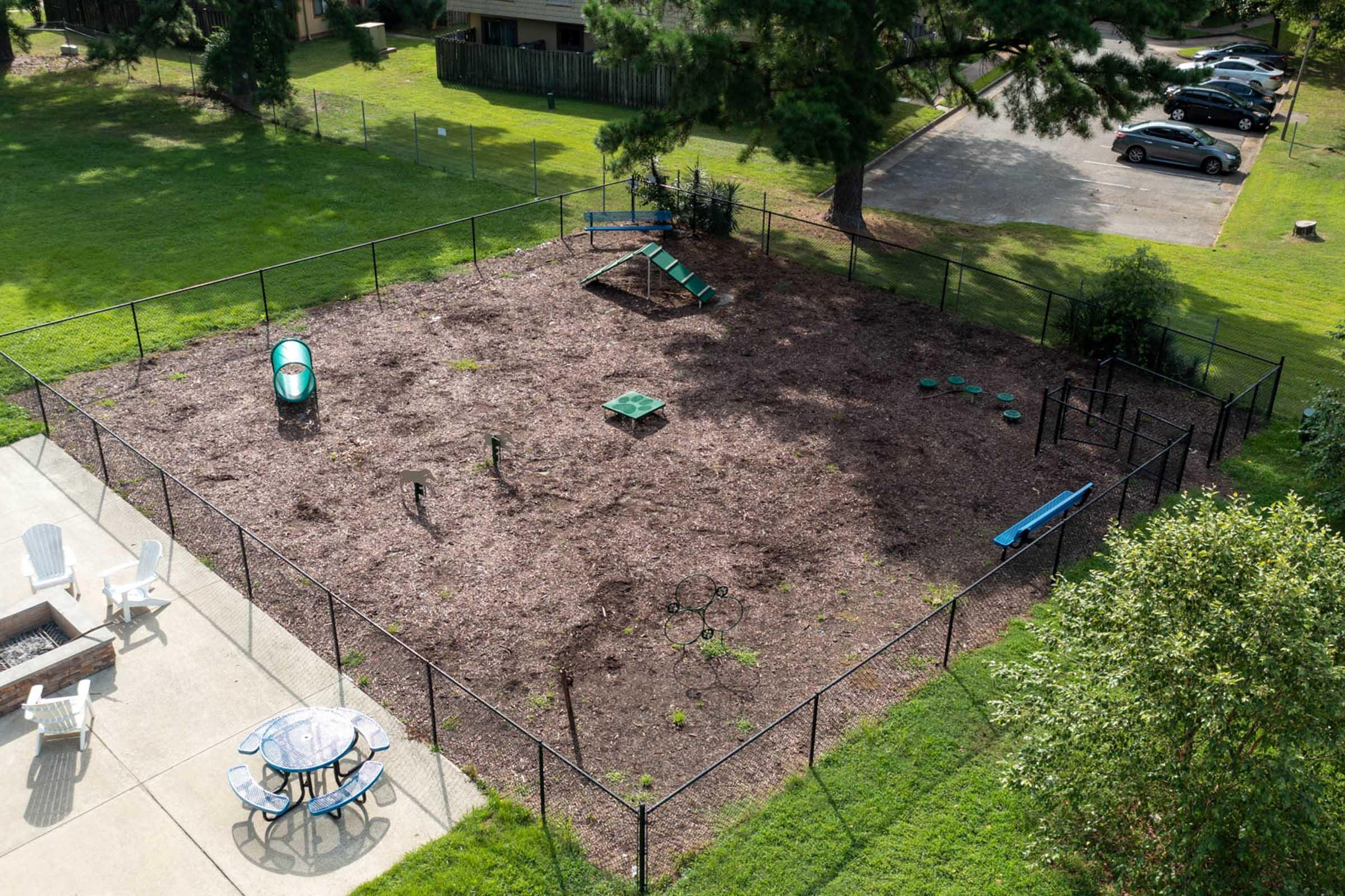Aerial view of a fenced-in playground area with wood mulch ground cover. The space includes a small slide, climbing structure, and various play equipment. Surrounding the playground are grassy areas and a nearby patio with a table and chairs. A few trees and a parking area are visible in the background.