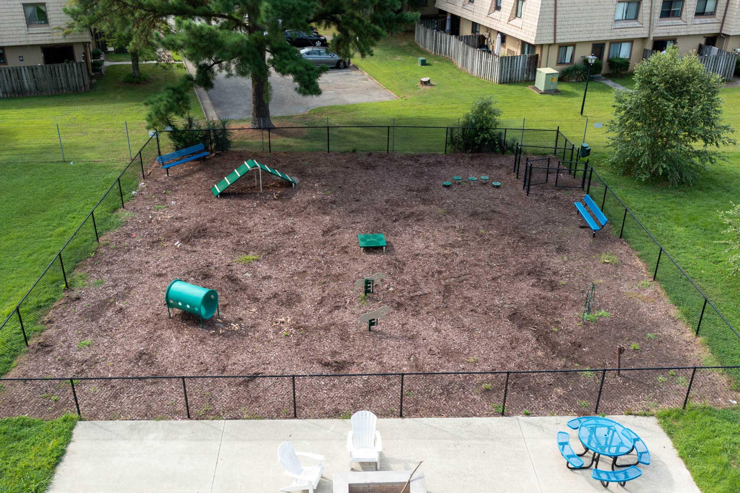 Aerial view of a fenced dog park with sparse ground cover, featuring a small agility structure (a ramp and tunnel), a few benches, and scattered green play equipment. Nearby, there are some trees and a grassy area, along with a couple of picnic tables visible in the foreground. The park is empty of dogs or people.