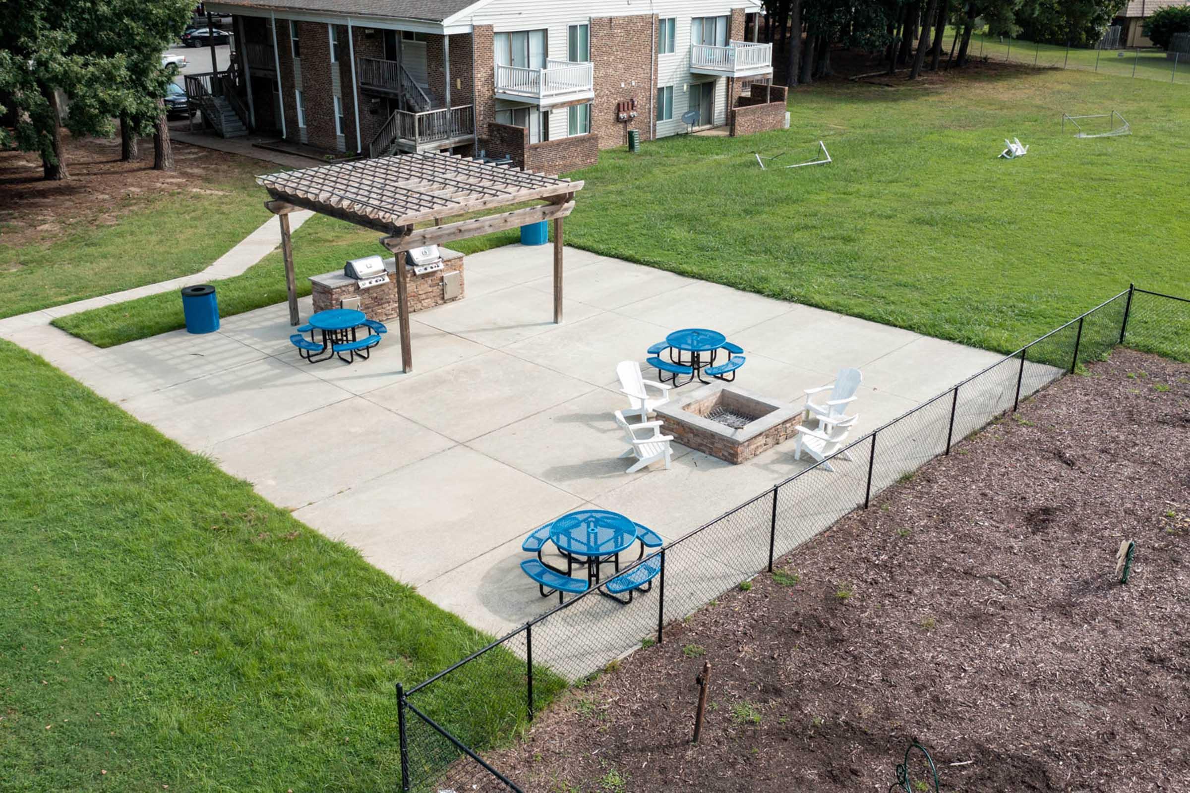 An outdoor recreational area featuring a concrete patio with blue picnic tables and white adirondack chairs. A shaded pergola with grills is present, alongside a grassy field and a fenced garden area. In the background, there are apartment buildings visible.