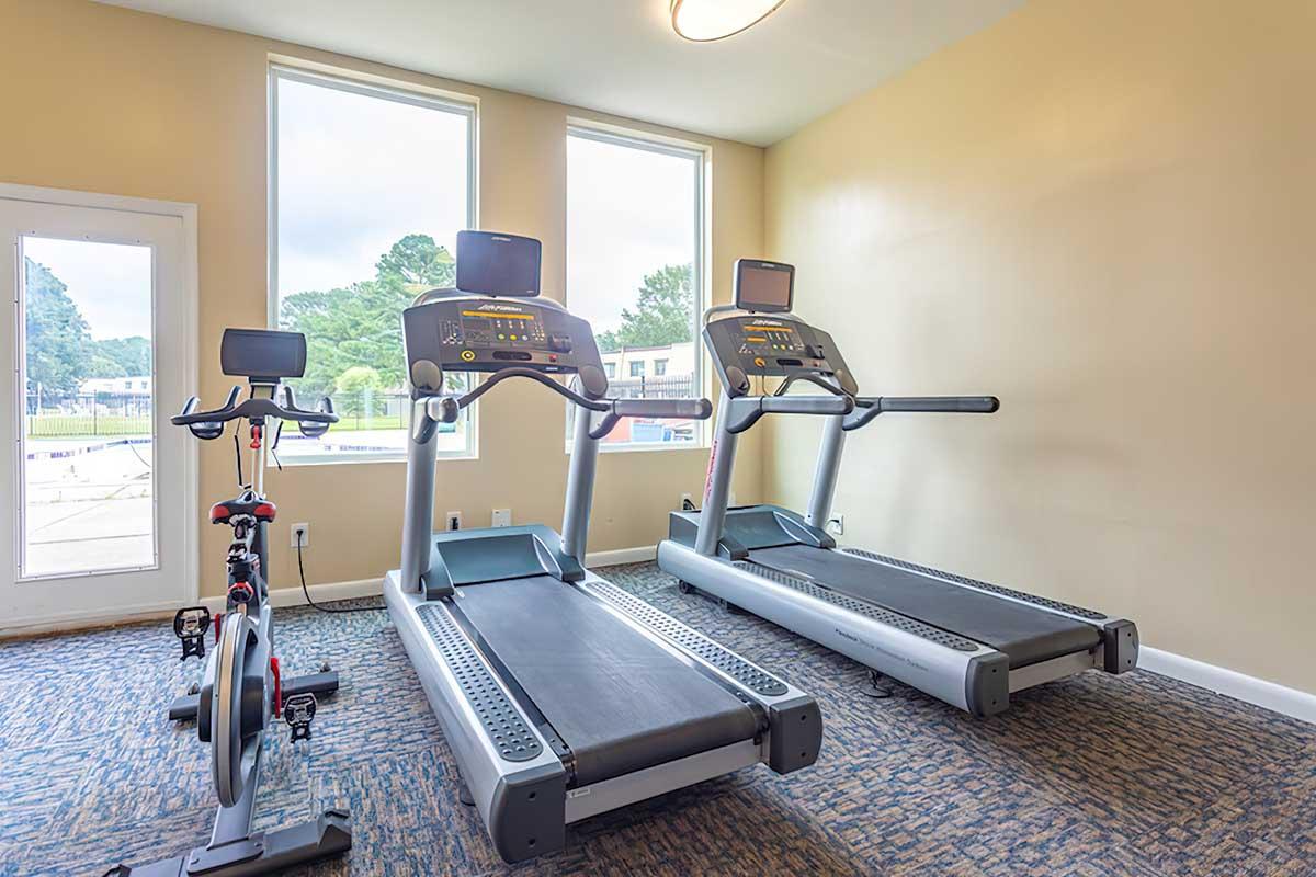 A fitness room featuring two treadmills and a stationary bike, with large windows providing natural light. The walls are a light color, and the flooring has a textured design. The equipment displays screens for workout tracking.