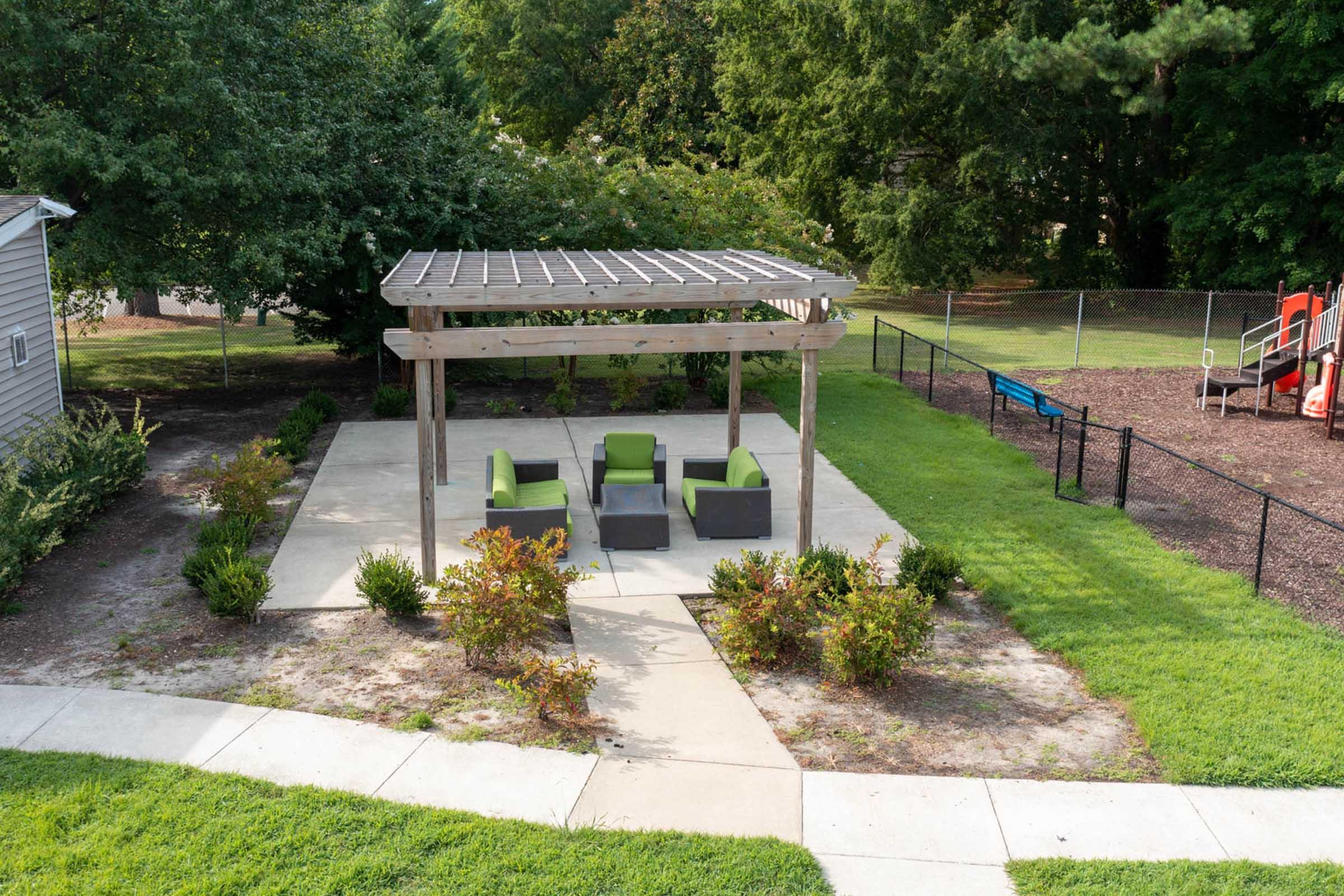 A landscaped backyard featuring a shaded seating area with four green chairs surrounding a small table. The seating is under a wooden pergola with a trellis. Surrounding the area are neatly trimmed shrubs and grass, along with a children's playground visible in the background.
