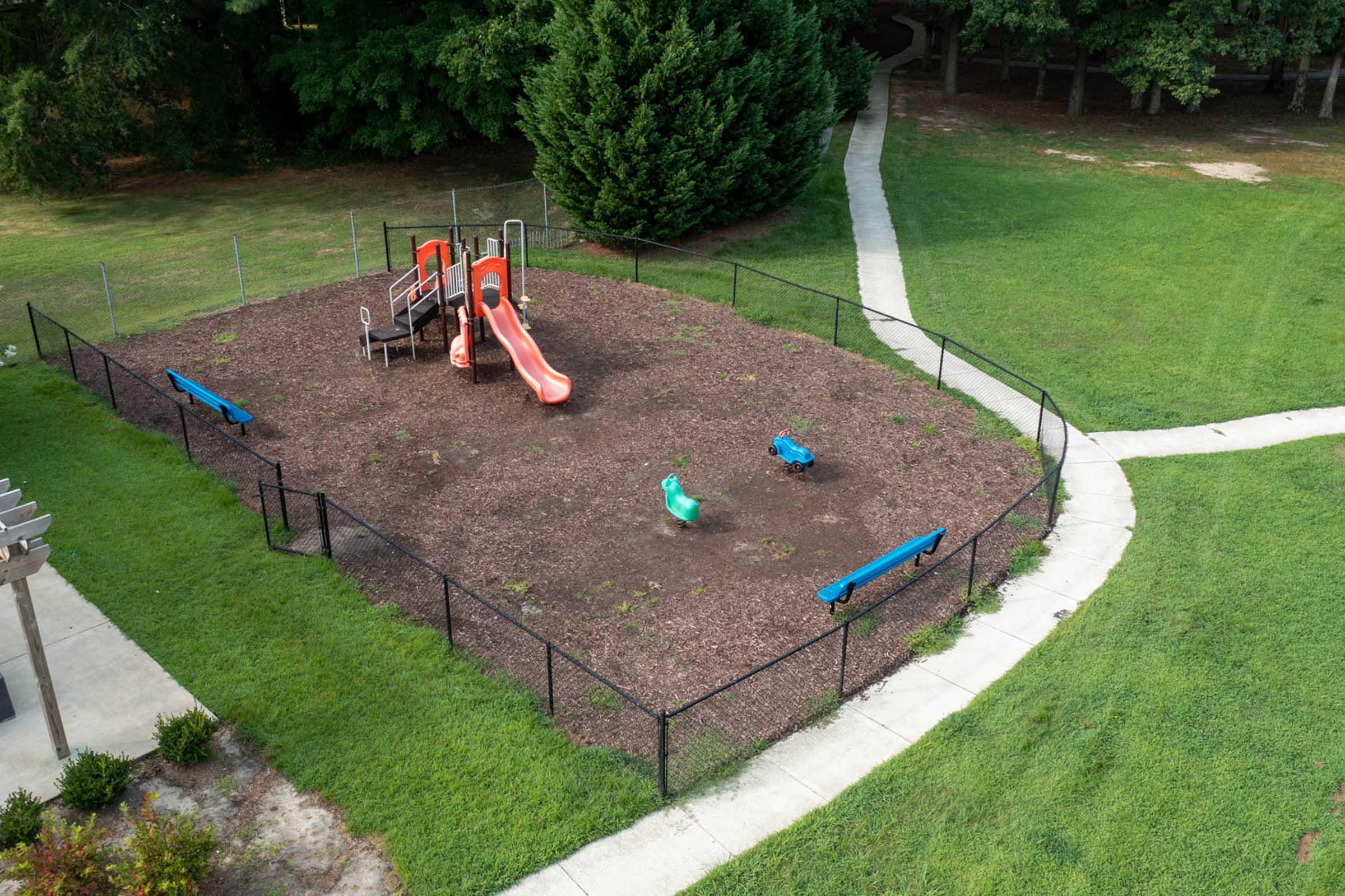 An aerial view of a playground with a red slide, two swings in blue and green, and a fenced grassy area. Pathways surround the playground, leading through green lawns and trees. The play area has mulch ground cover and appears to be unoccupied.