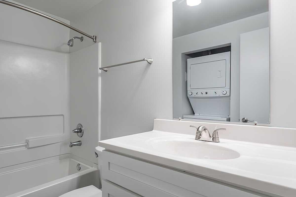 A modern bathroom featuring a white bathtub, shower area, and a simple vanity with a sink. A mirror is mounted above the sink, and a stacked washer and dryer unit is visible in a recessed space. The walls and surfaces are painted in light colors, creating a clean and minimalist look.