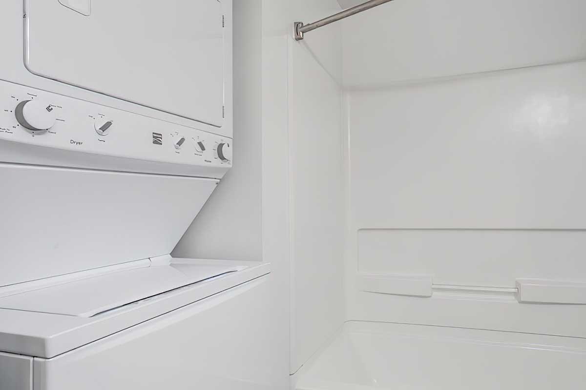 A white stacked washer and dryer unit positioned next to a clean, empty shower space with a shower rod. The walls and fixtures are white, giving a bright and spacious appearance.