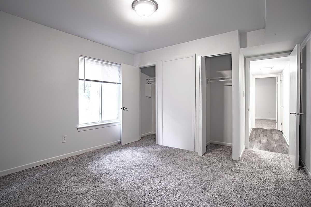 Empty room featuring light gray walls and carpeting, with a window allowing natural light. Two closets with sliding doors are visible on one side. The space is clean and minimalistic, with a ceiling light fixture and an adjacent door leading to another area.