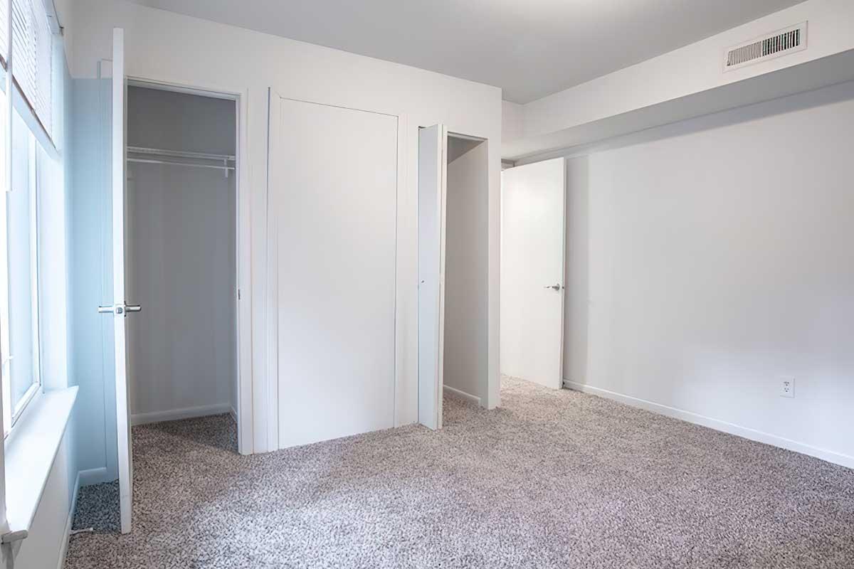 Empty room featuring light-colored walls and plush carpet. There are two open doors leading to closets and another door on the right side. The room is well-lit with natural light coming from a window, creating a spacious and clean aesthetic.