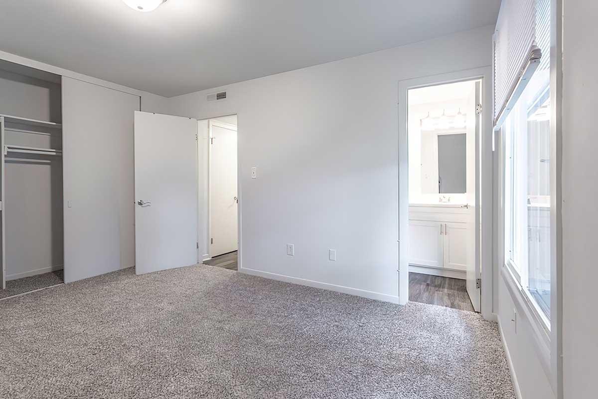 A spacious, empty room with light gray carpet, featuring an open closet on the left, a white door leading to a bathroom on the right, and a window allowing natural light. The walls are painted in a neutral shade, creating a serene atmosphere.