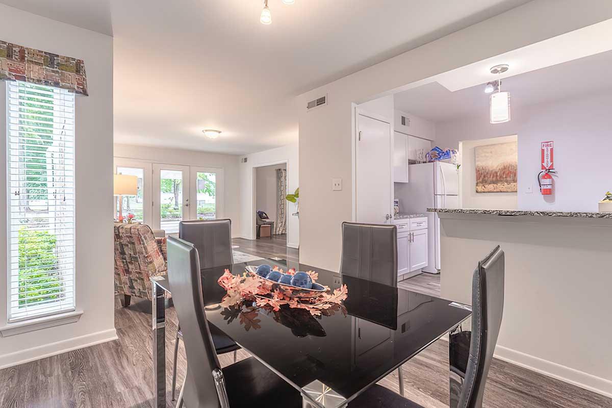 A modern dining area featuring a sleek black table with a decorative centerpiece of leaves and fruit. In the background, there are large windows allowing natural light, a well-equipped kitchen with white cabinetry, and inviting living spaces with contemporary furnishings.