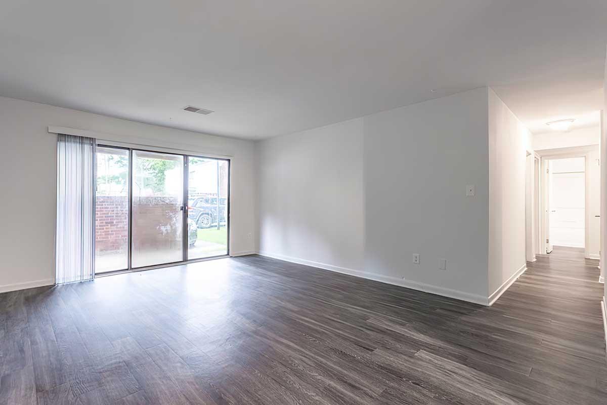 Spacious empty living room with hardwood-style flooring, featuring a large sliding glass door leading to an outdoor space. Light-colored walls create an airy atmosphere. A hallway is visible on the right, leading to additional rooms. Natural light fills the space.