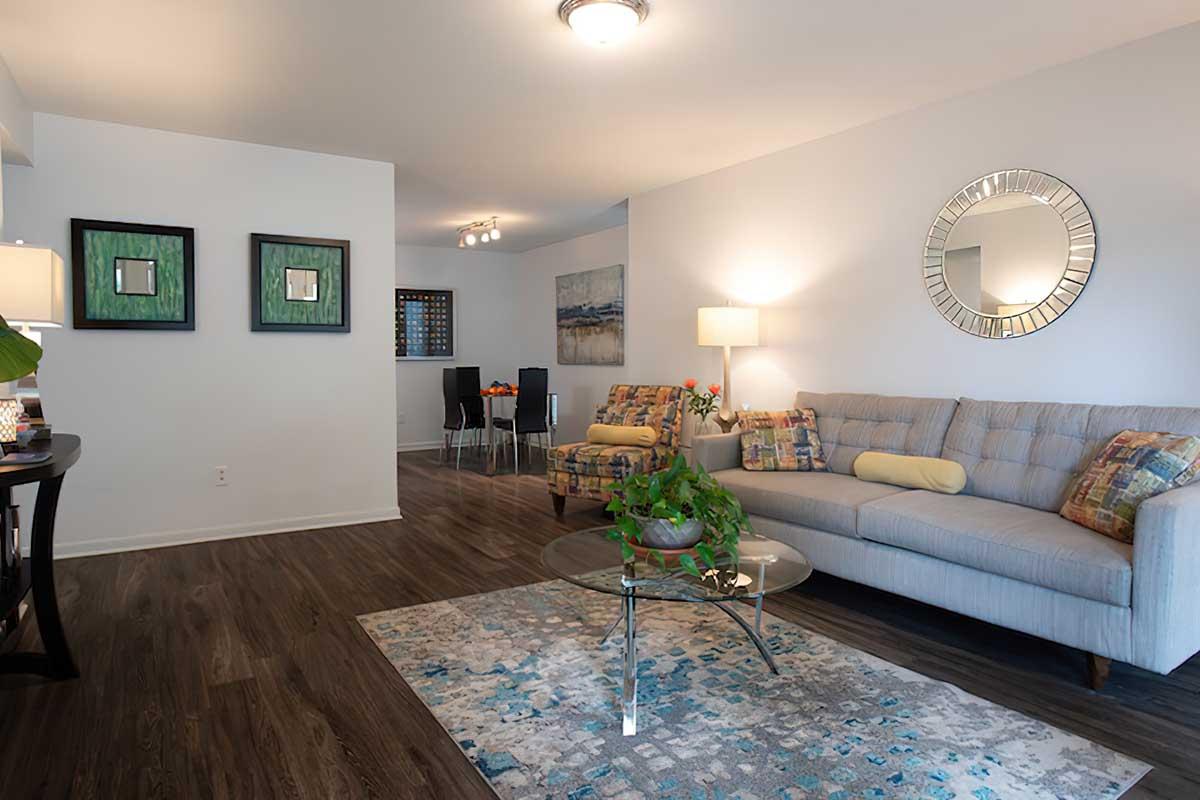 A modern living room featuring a light gray sofa with colorful cushions, a glass coffee table with a potted plant, and a decorative area rug. The walls are painted white, adorned with two framed artworks, and there are lamps providing soft lighting. In the background, a dining area is visible with black chairs and a table.