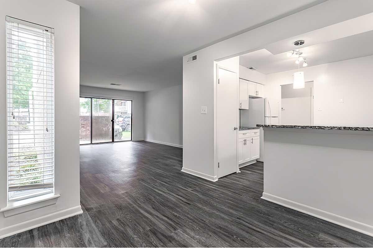 A modern, empty apartment interior featuring an open layout. The living area has large windows with blinds and sleek flooring. A kitchen area with white cabinets and granite countertops is visible, along with a view of a sliding glass door leading to an outdoor space. The walls are painted light, creating a bright atmosphere.