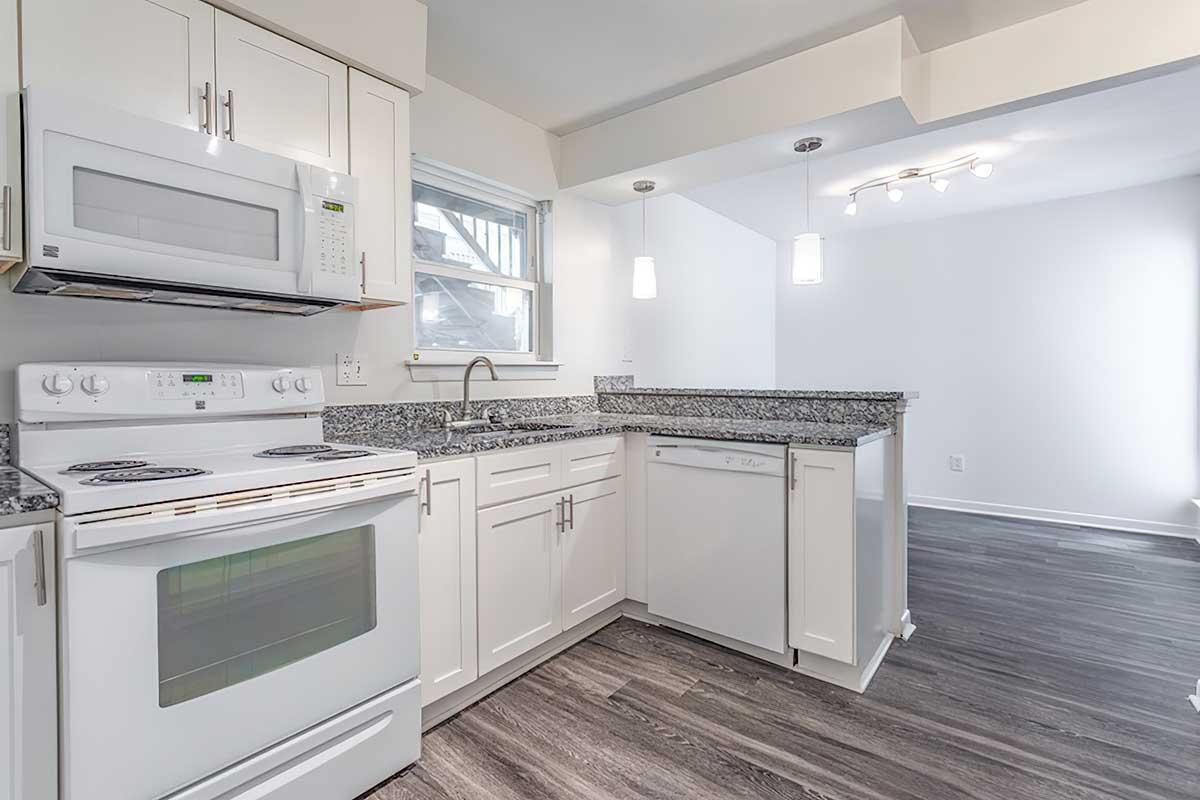Modern kitchen featuring white cabinets, granite countertops, a built-in oven and microwave, a dishwasher, and pendant lighting. The space has a light color palette with a window for natural light and gray hardwood flooring.