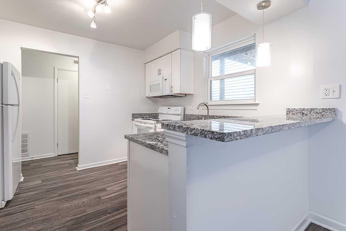 A modern kitchen featuring white cabinetry, granite countertops, and stainless steel appliances. The space is well-lit with pendant lights and a window providing natural light. A doorway leads to a separate area, and the floor is covered with dark wood laminate.