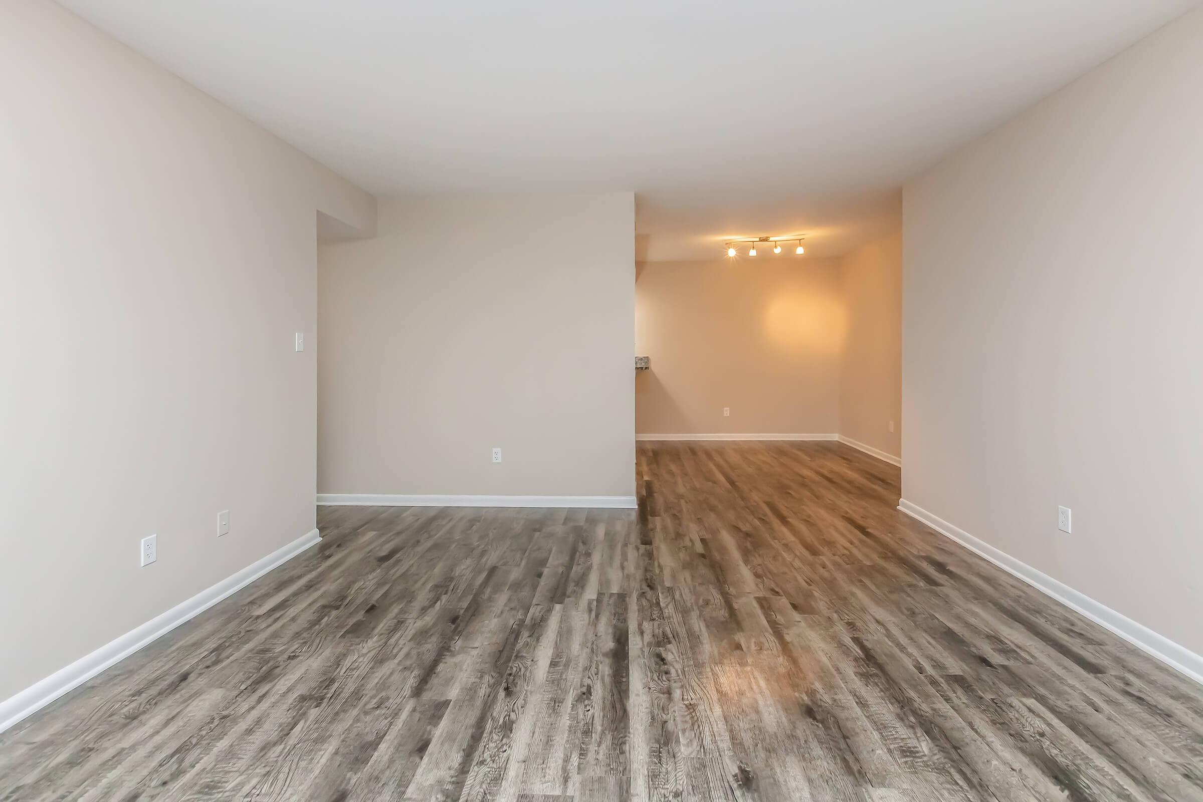 Empty interior of a modern living space featuring light-colored walls and a wood-like floor. Natural light from a ceiling fixture illuminates the room. The layout is open, transitioning to an adjacent area with a faint outline of a kitchen or dining space in the background.