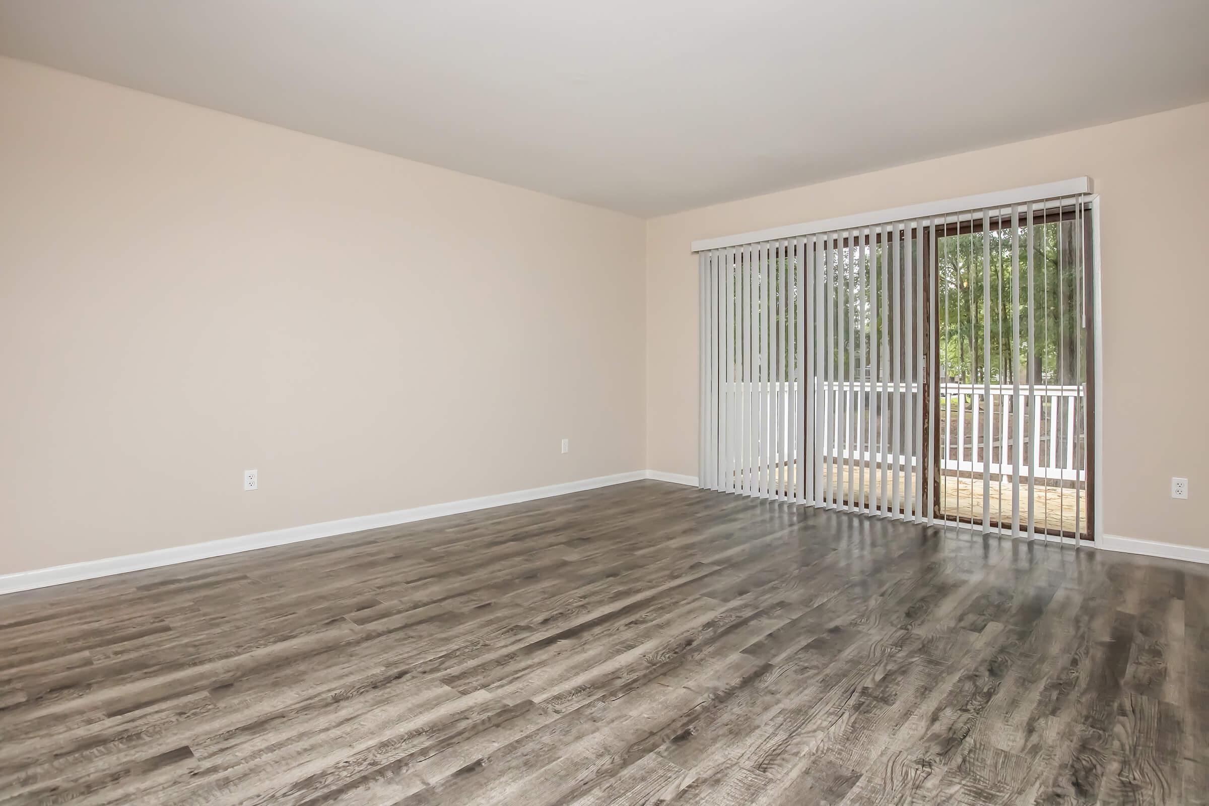 A spacious, empty room with light beige walls and a large sliding glass door featuring vertical blinds. The floor is covered in wood-like laminate, creating a modern aesthetic. Natural light streams in from outside, illuminating the space.