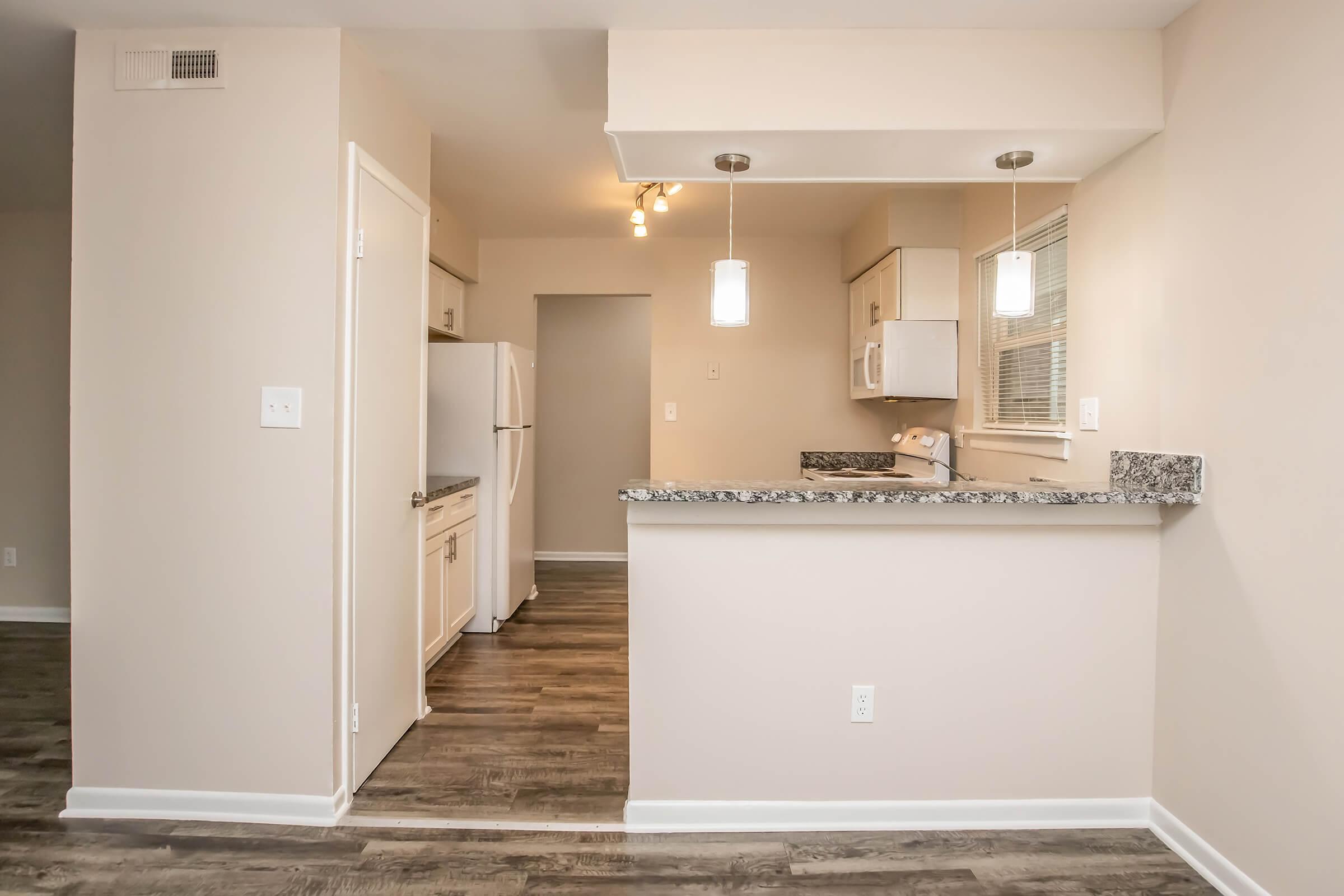 A view of a modern kitchen featuring light-colored walls, a granite countertop with pendant lighting, and white cabinetry. The space includes a refrigerator, microwave, and an open layout leading to a living area with wooden flooring.