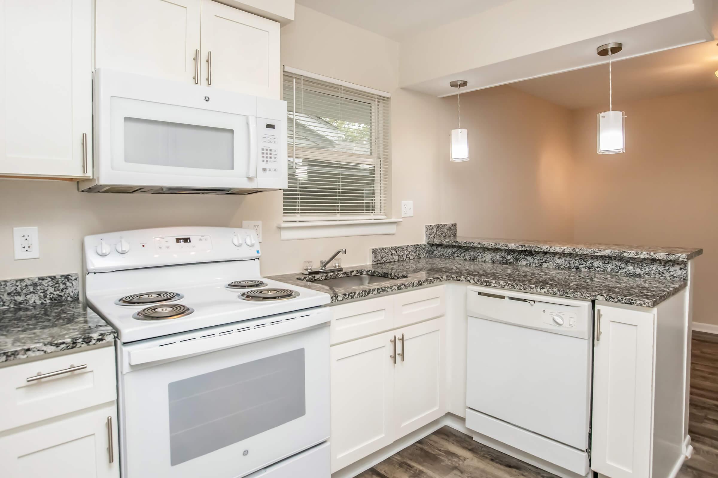 Modern kitchen featuring white cabinetry, granite countertops, and stainless steel appliances. Includes a white stove and microwave, a dishwasher, and pendant lighting above the countertop. Natural light comes through a window, and there is a seamless transition to the adjacent dining area.